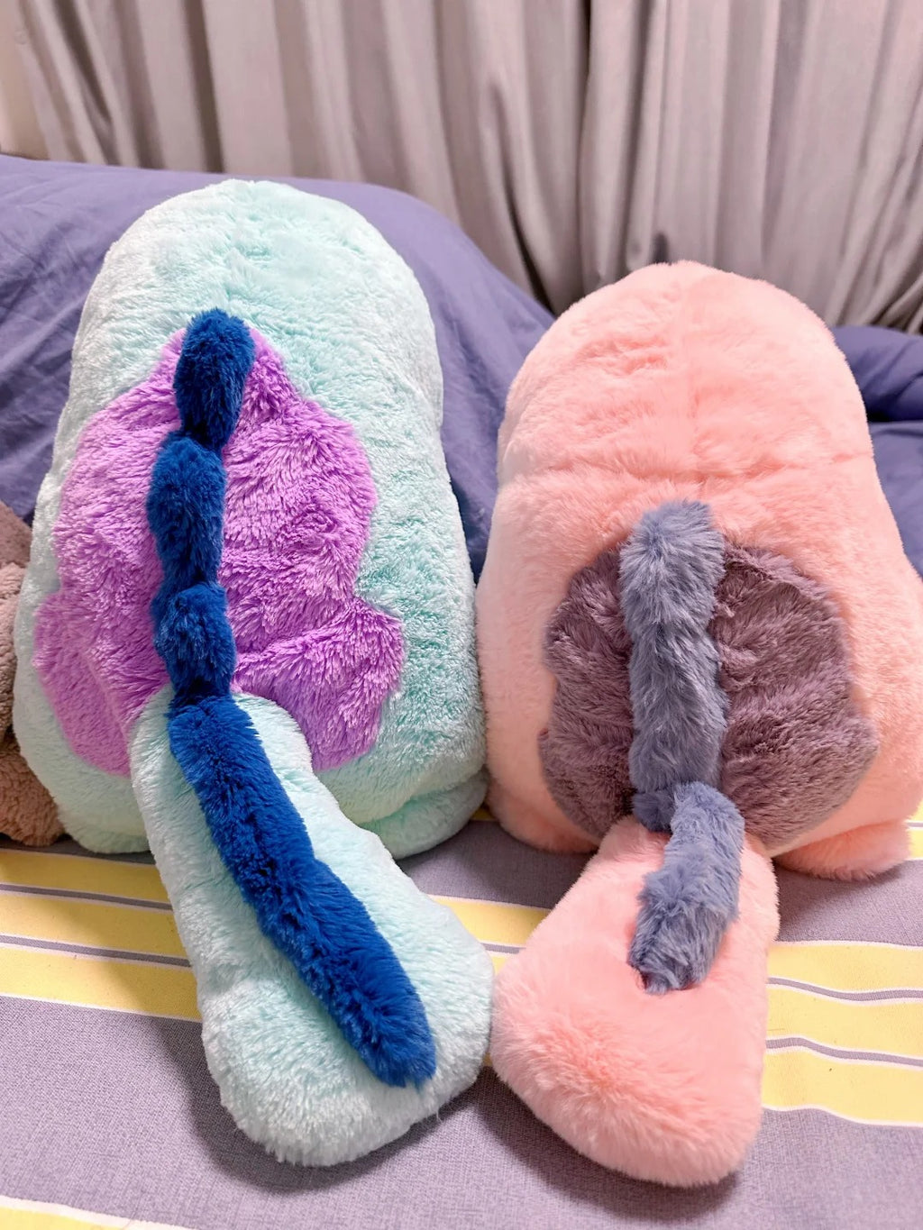 Two  Quagsire  plush animal toys with fluffy textures, one mint green with a purple and blue tail-like detail, and one peach pink with a gray and pink tail-like detail, resting on a bed.