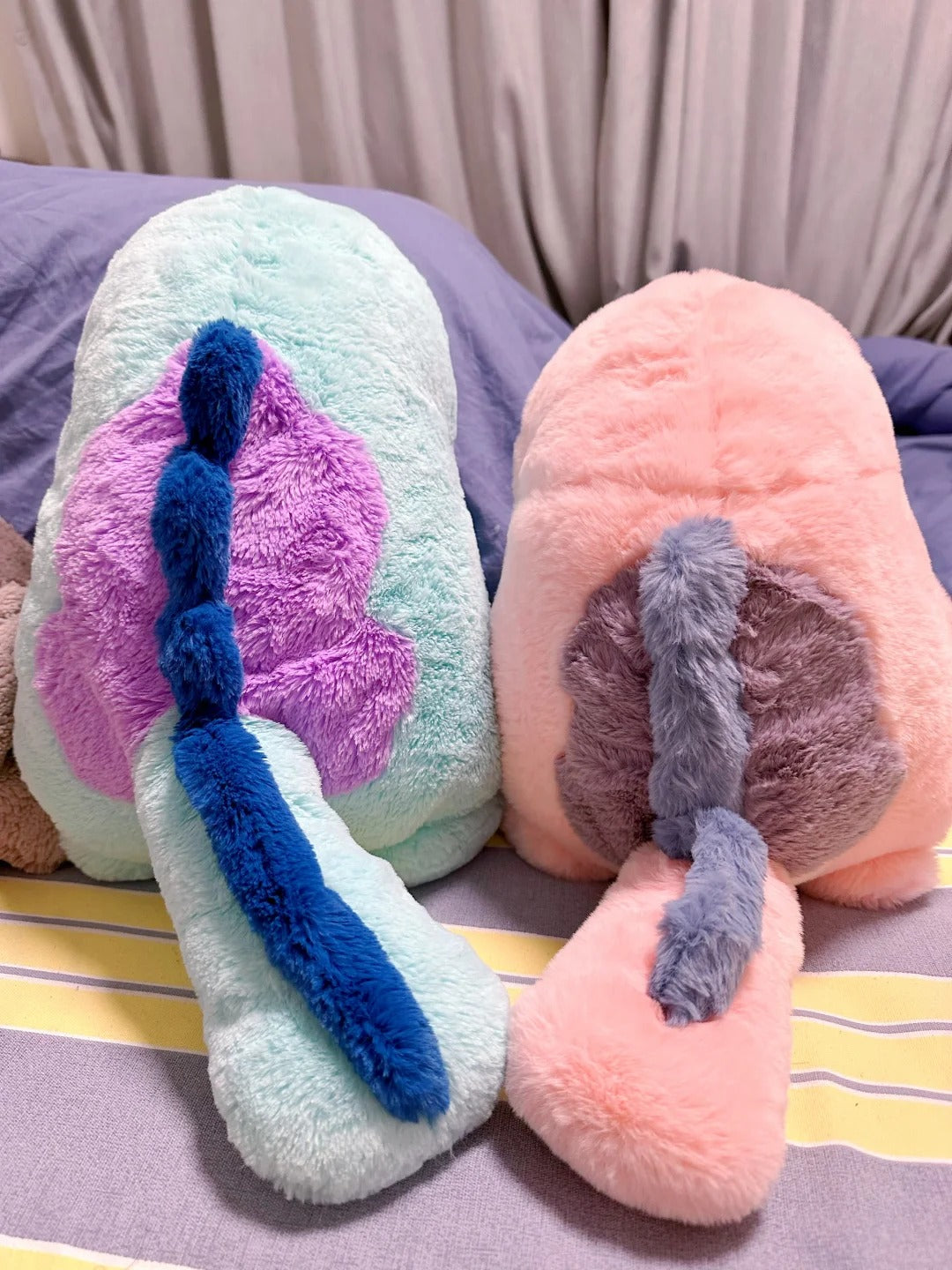 Two  Quagsire  plush animal toys with fluffy textures, one mint green with a purple and blue tail-like detail, and one peach pink with a gray and pink tail-like detail, resting on a bed.
