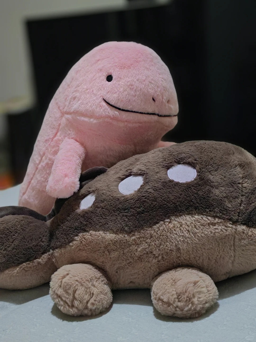 Two plush toys, a pink  Quagsire  whale-shaped plush standing behind a brown and beige fish-shaped plush, set against a dark blurred background.