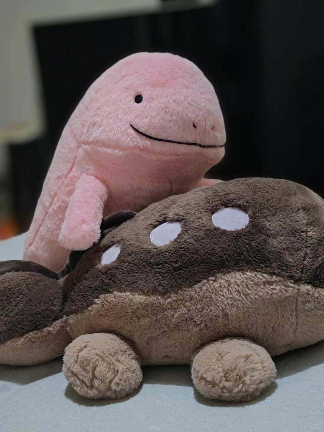 Two plush toys, a pink  Quagsire  whale-shaped plush standing behind a brown and beige fish-shaped plush, set against a dark blurred background.