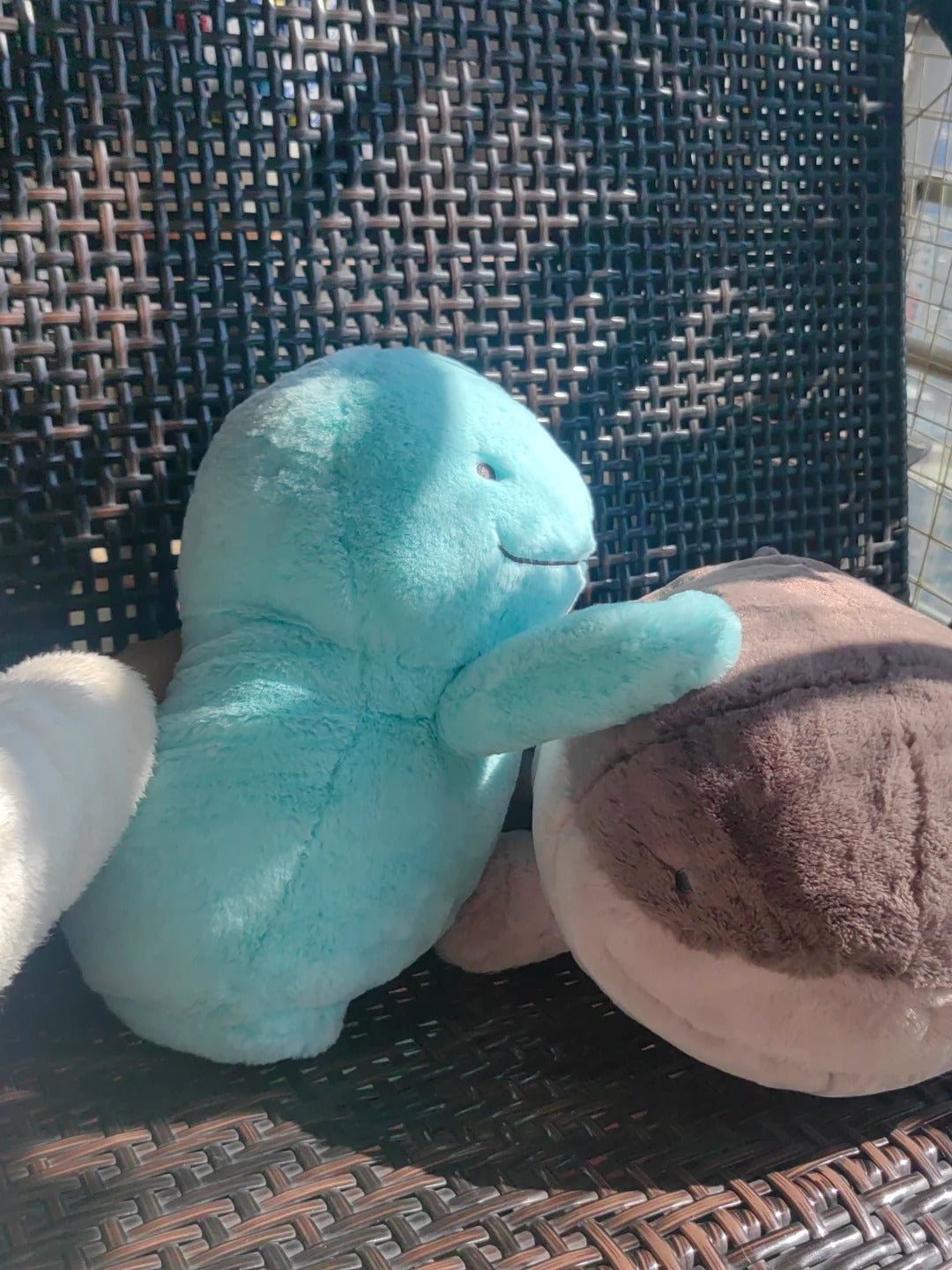 A light blue Quagsire plush toy sitting on a dark brown woven outdoor chair next to a brown and beige stuffed animal.