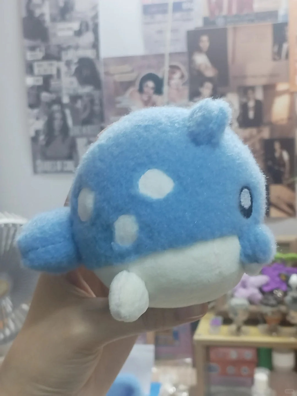 A blue and white Spheal plush toy shaped like a cartoon fish or whale, held in a person's hand in a room with posters and shelves in the background.