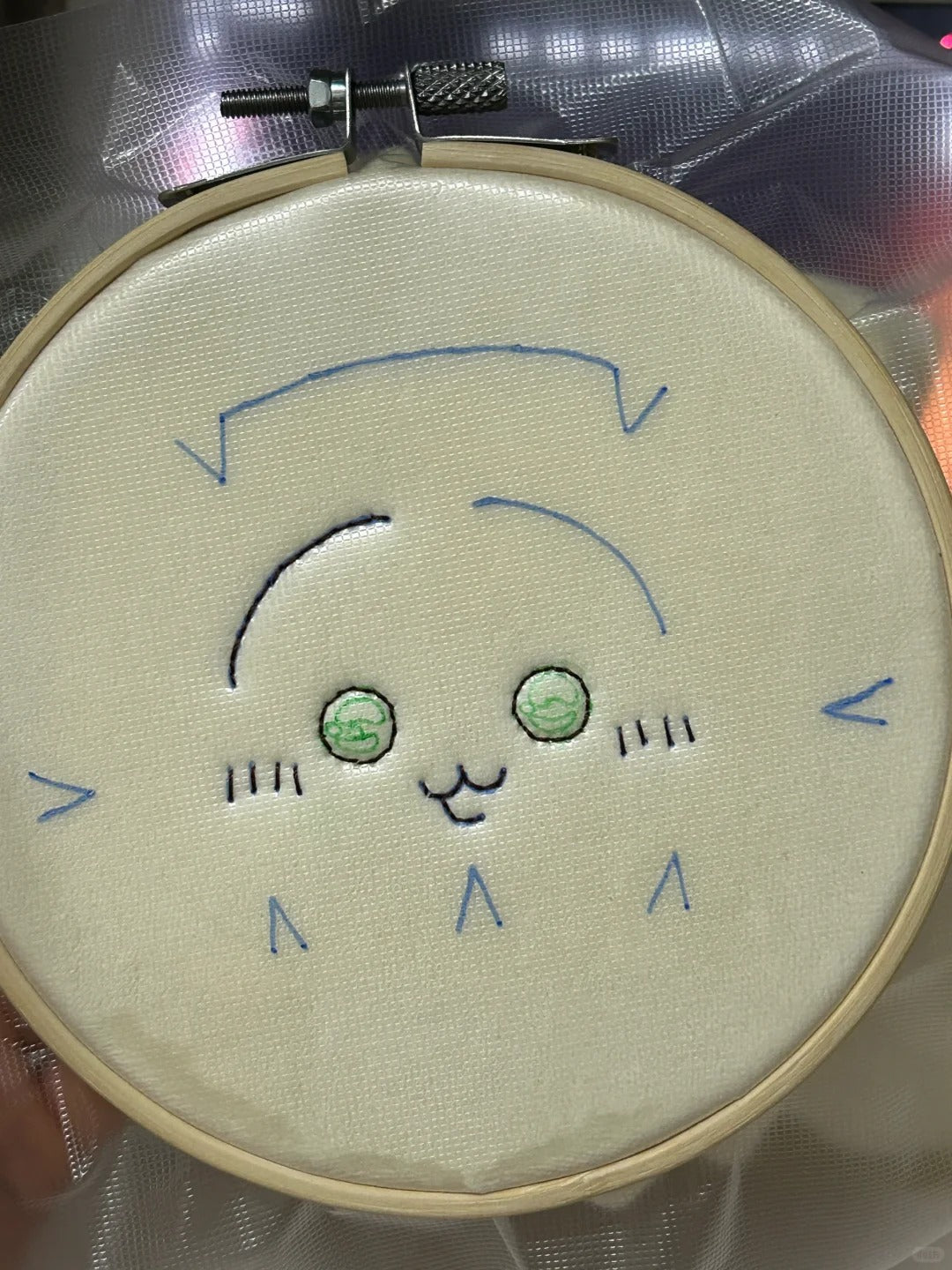 A close-up of a circular embroidery hoop holding off-white fabric with a partially stitched cartoon face. The embroidery includes blue outline details, green embroidered eyes, and simple facial features.