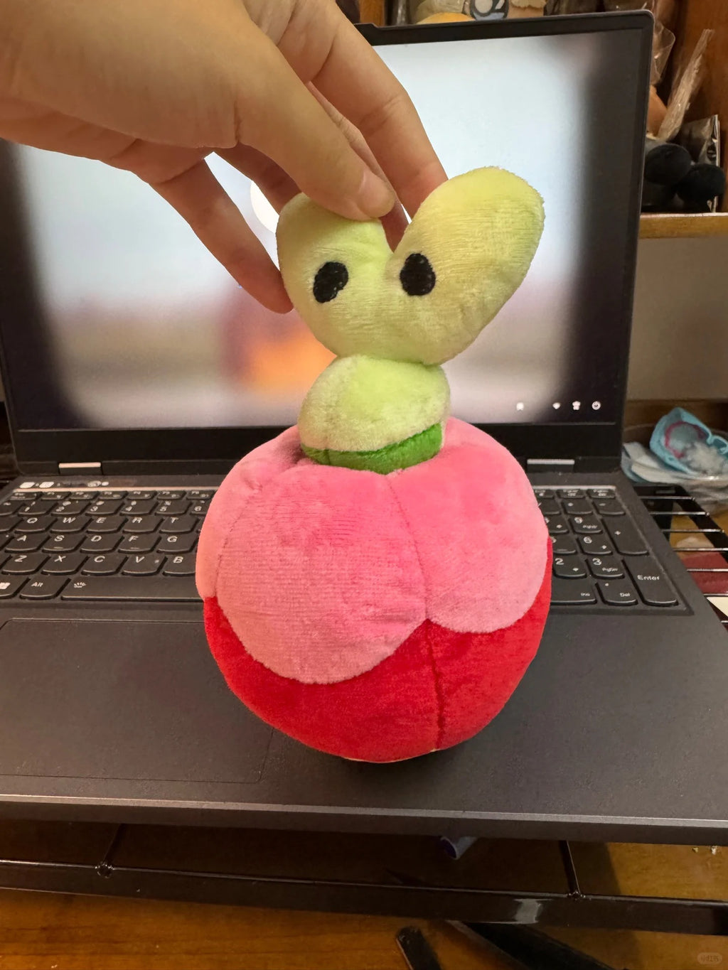 A Applin plush toy shaped like a stylized fruit or flower with a pink and red round body, a light green stem, and a pale yellow heart-shaped top with black eyes, being held by a hand in front of an open laptop.
