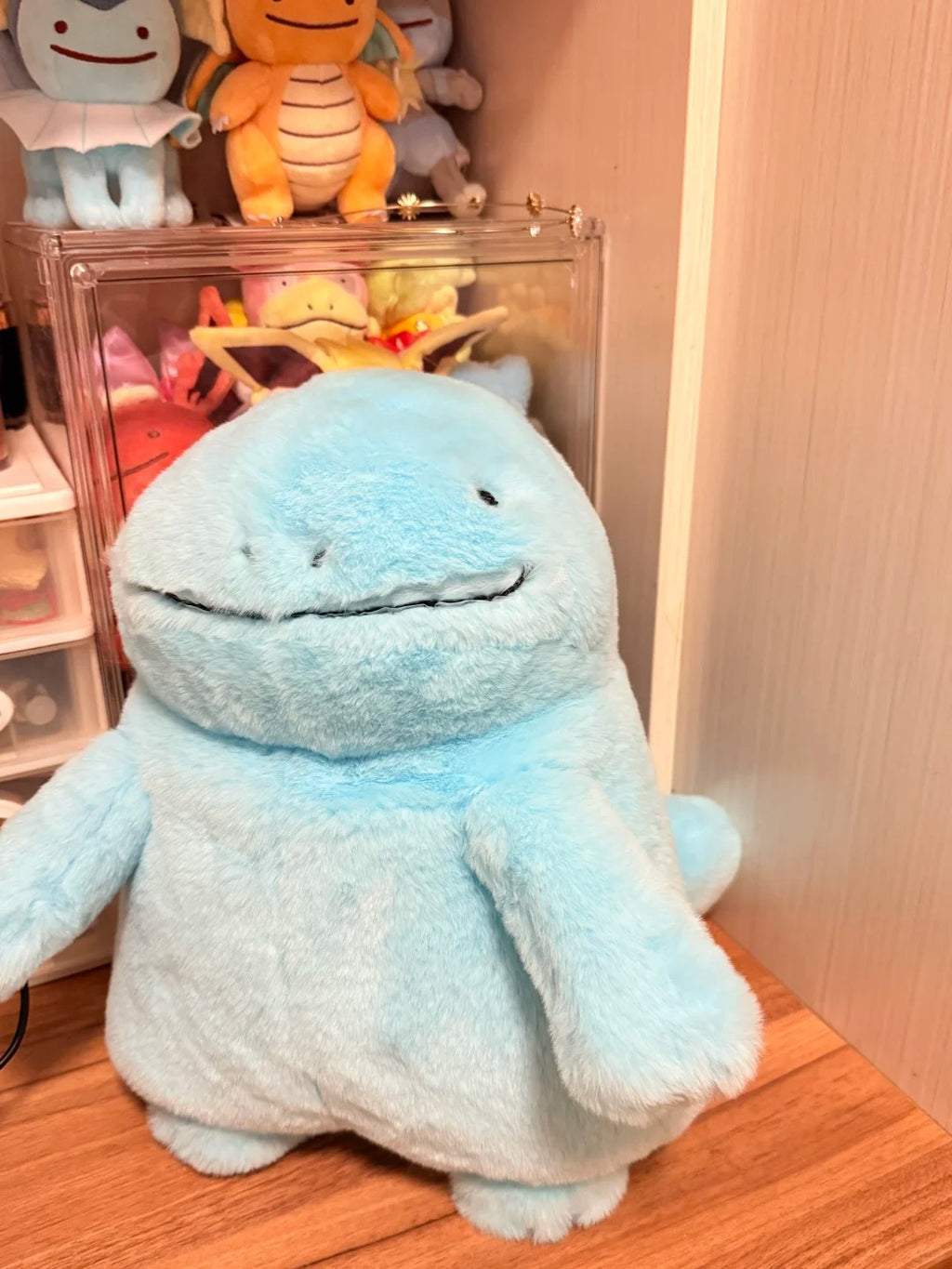 A light blue Quagsire plush toy resembling a cartoon amphibious creature, photographed standing on a wooden surface in front of a shelf with other stuffed animals and a clear display case.