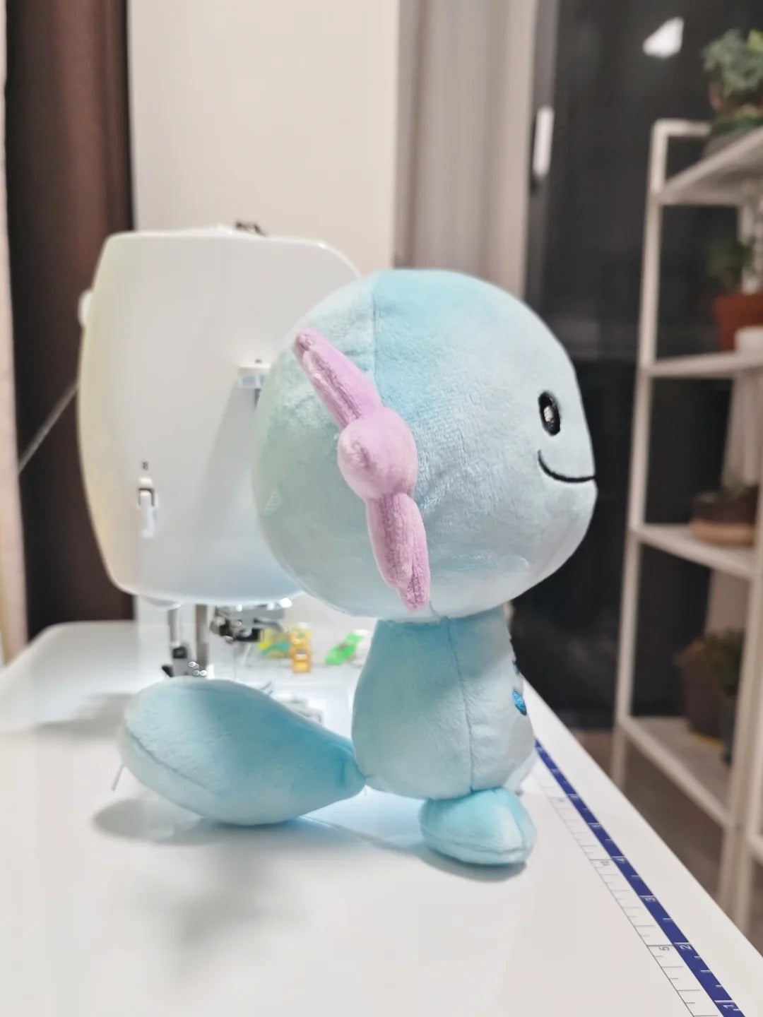 A light blue Wooper plush toy with a pink bow on its head, sitting on a white sewing table in front of a white sewing machine, with a blurred indoor room in the background.