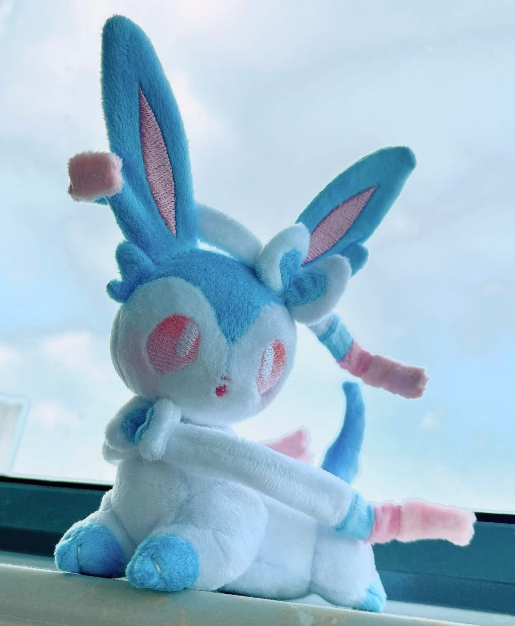 A blue and white Sylveon plush toy with pink accents, resembling a cute fantasy creature, sitting on a windowsill with a bright sky visible in the background.