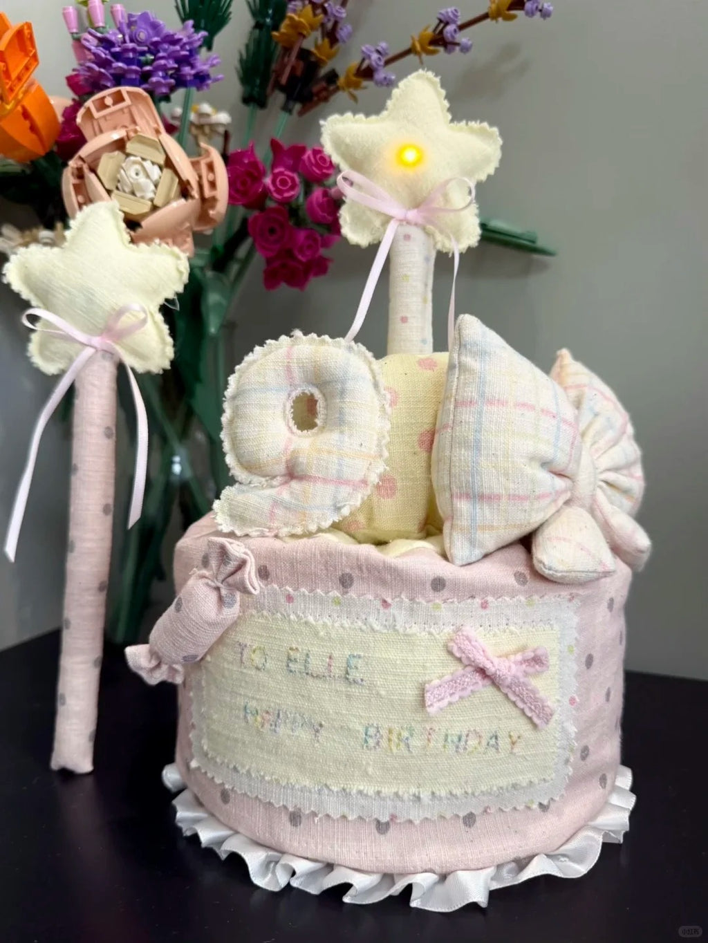 A handmade baby shower diaper cake decoration set in pastel pink and cream, featuring a ruffled diaper base, embroidered 'TO ELLE 10TH BIRTHDAY' text, plush pillows, and two star-shaped baby towers with small built-in lights, displayed on a dark surface with flowers in the background.