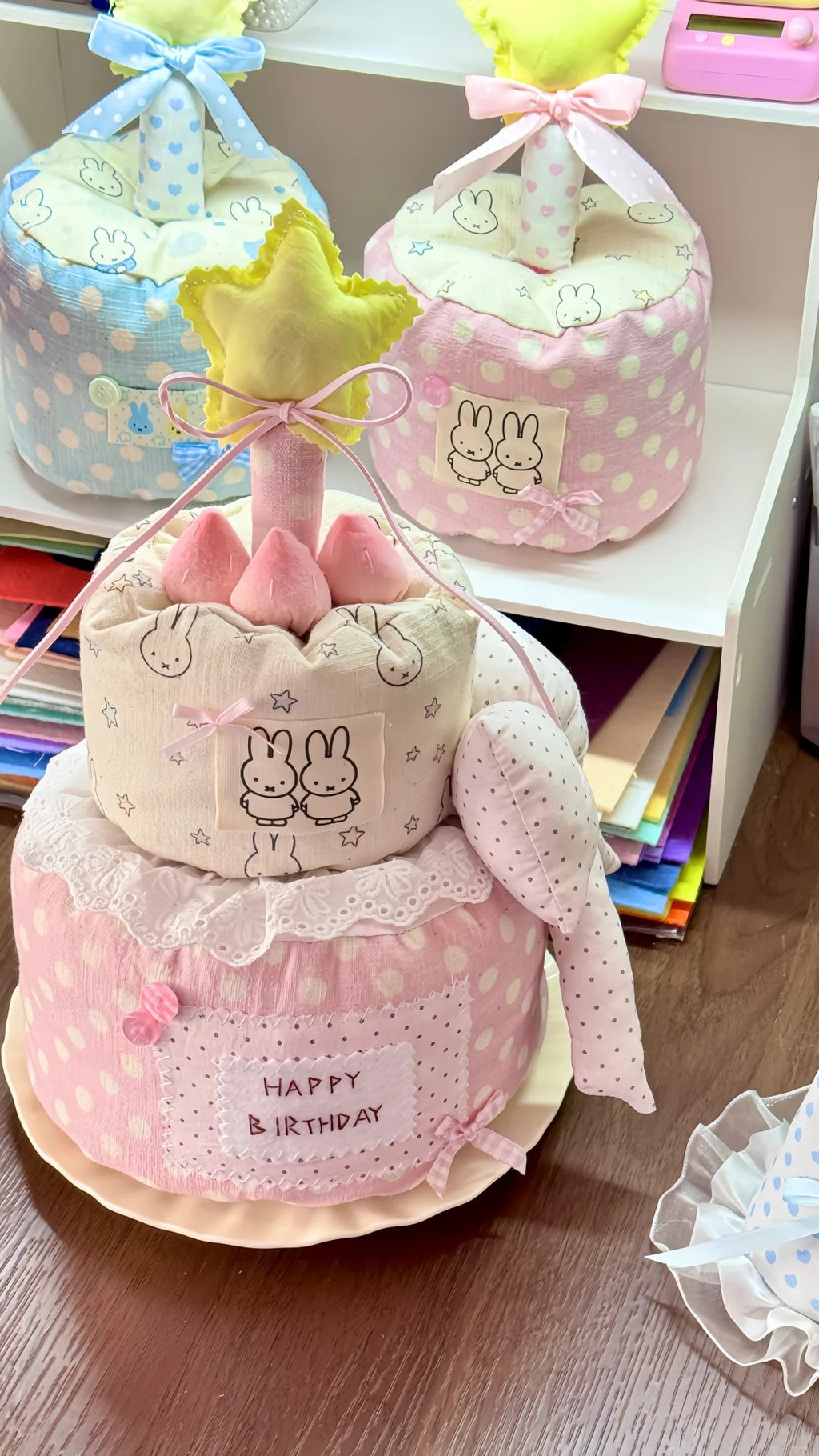 A handmade fabric cake-shaped baby shower favor or gift container in pink and white polka dots, decorated with bunny graphics, lace trim, a yellow star topper, and a 'HAPPY BIRTHDAY' sign. Two similar cake-shaped favors in blue and pink are visible in the background on a white shelf.