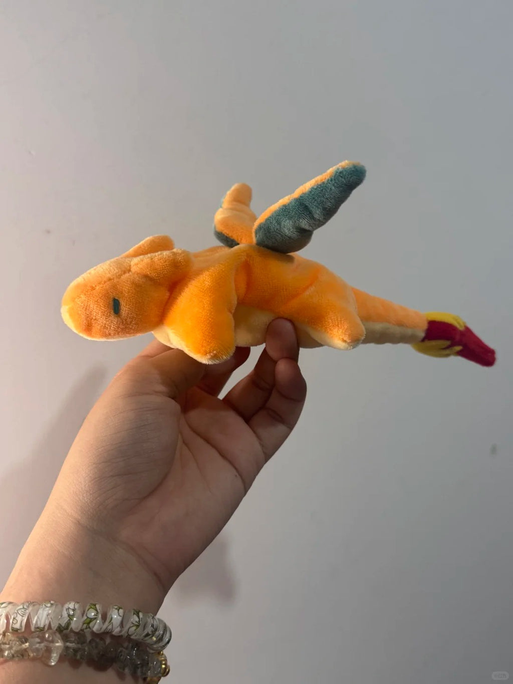 An orange dragon plush toy dragon with teal wings and a pink tail, held in a person's hand against a plain light gray wall.