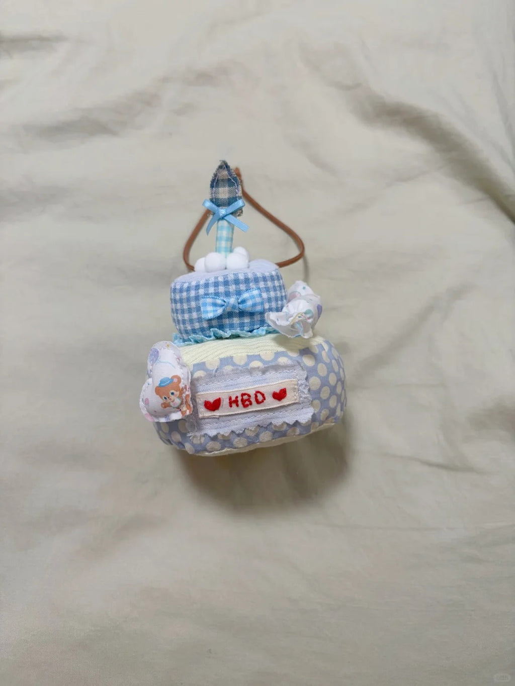 A handmade baby diaper bag charm shaped like a tiered cake, decorated with blue and white gingham patterns, a blue ribbon bow, a small toy airplane on top, and a white patch with red hearts and the text 'HBD'.