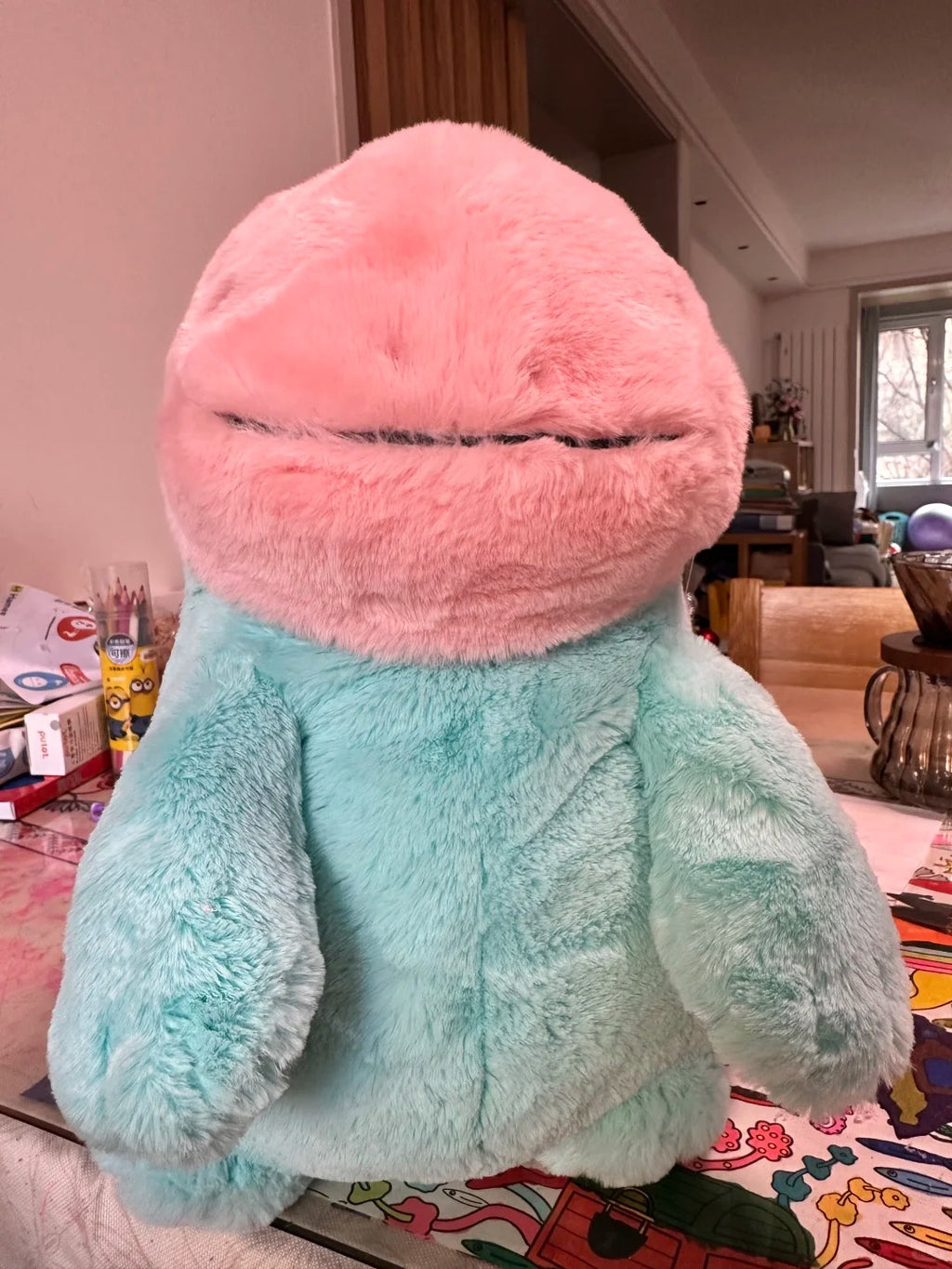 A Quagsire plush toy shaped like a round, soft creature with a pink fuzzy head and a mint green fuzzy body, photographed indoors on a patterned surface with a living room background.