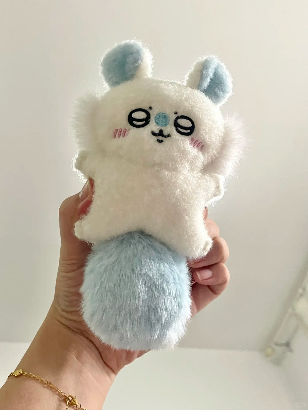 A hand holding a small handmade momonga plush character toy with a fluffy white body, light blue ears and tail, and embroidered closed eyes and pink cheeks against a plain light background.