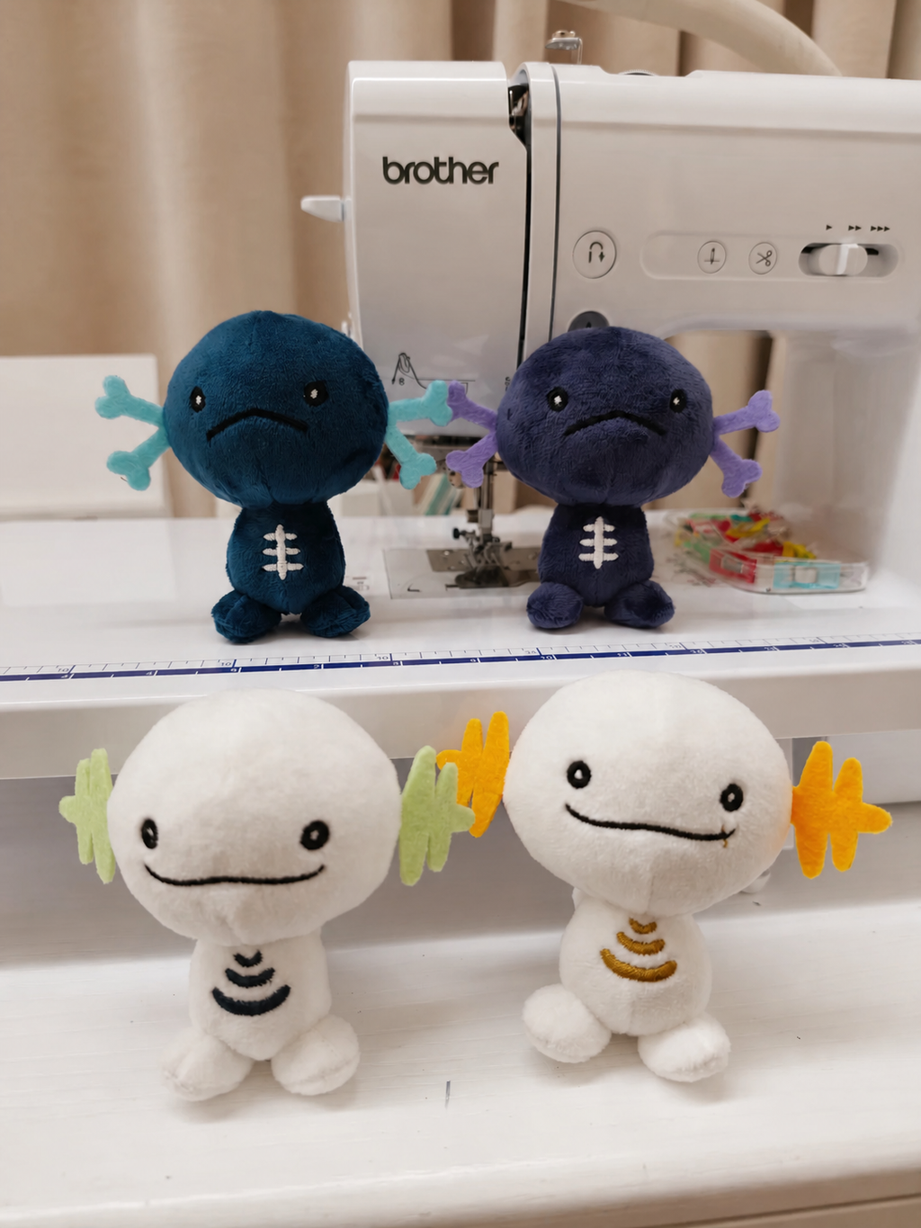 Four Wooper small plush toy figures arranged on a white sewing machine in a home setting. The top pair are dark blue and purple with light blue and purple leaf-like accents, and the bottom pair are white with black and gold accents.