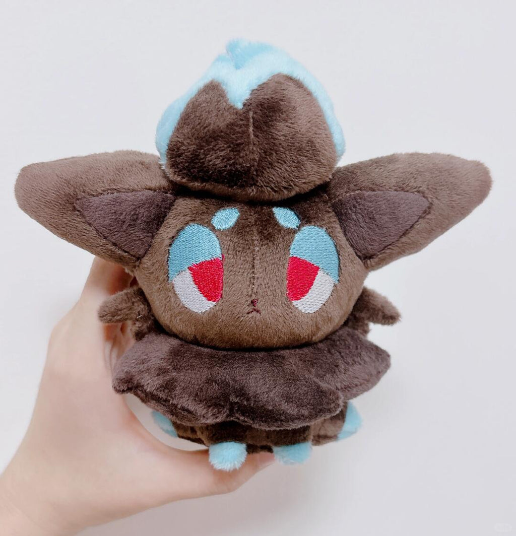 A small zorua plush toy held in a human hand against a plain light background. The toy is brown with light blue accents on its head and feet, and large embroidered eyes with red and white details.