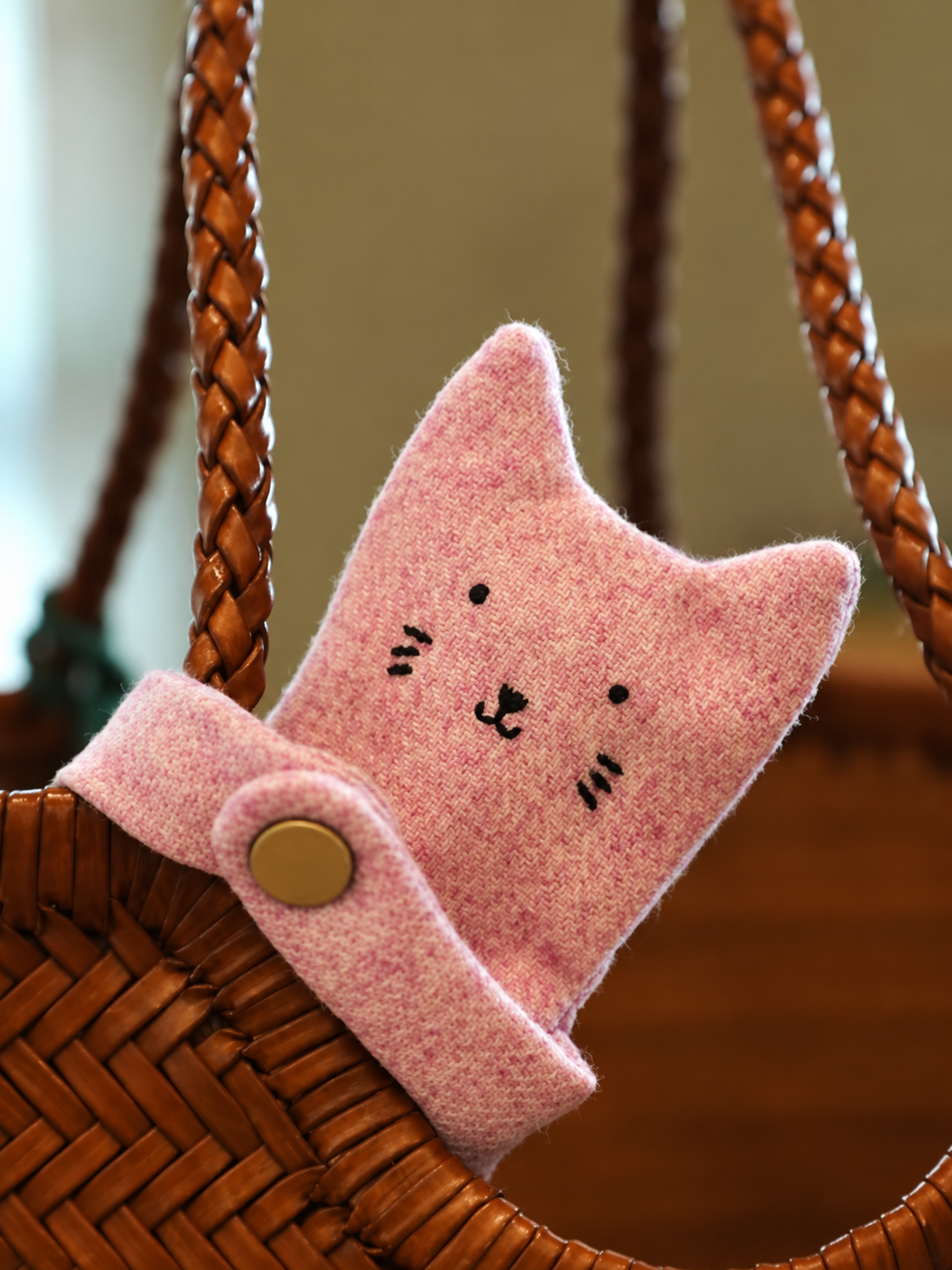 A pink felt cat-shaped accessory with embroidered facial features and a snap closure, attached to a brown woven handbag with braided straps.