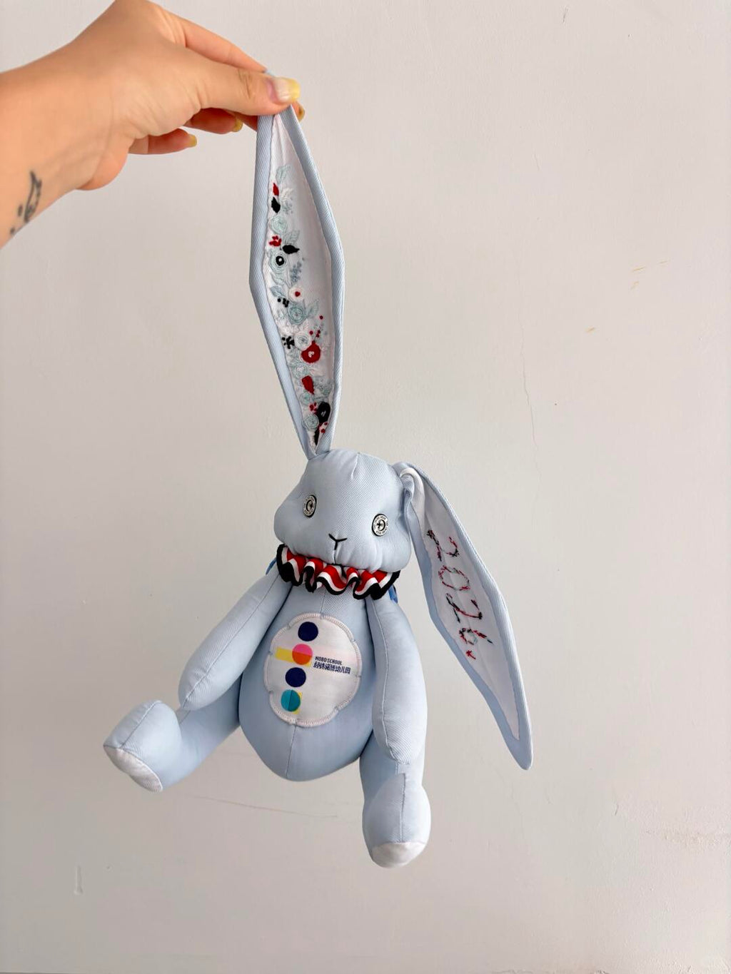 Light blue denim bunny plush hanging by one embroidered ear against a plain wall.