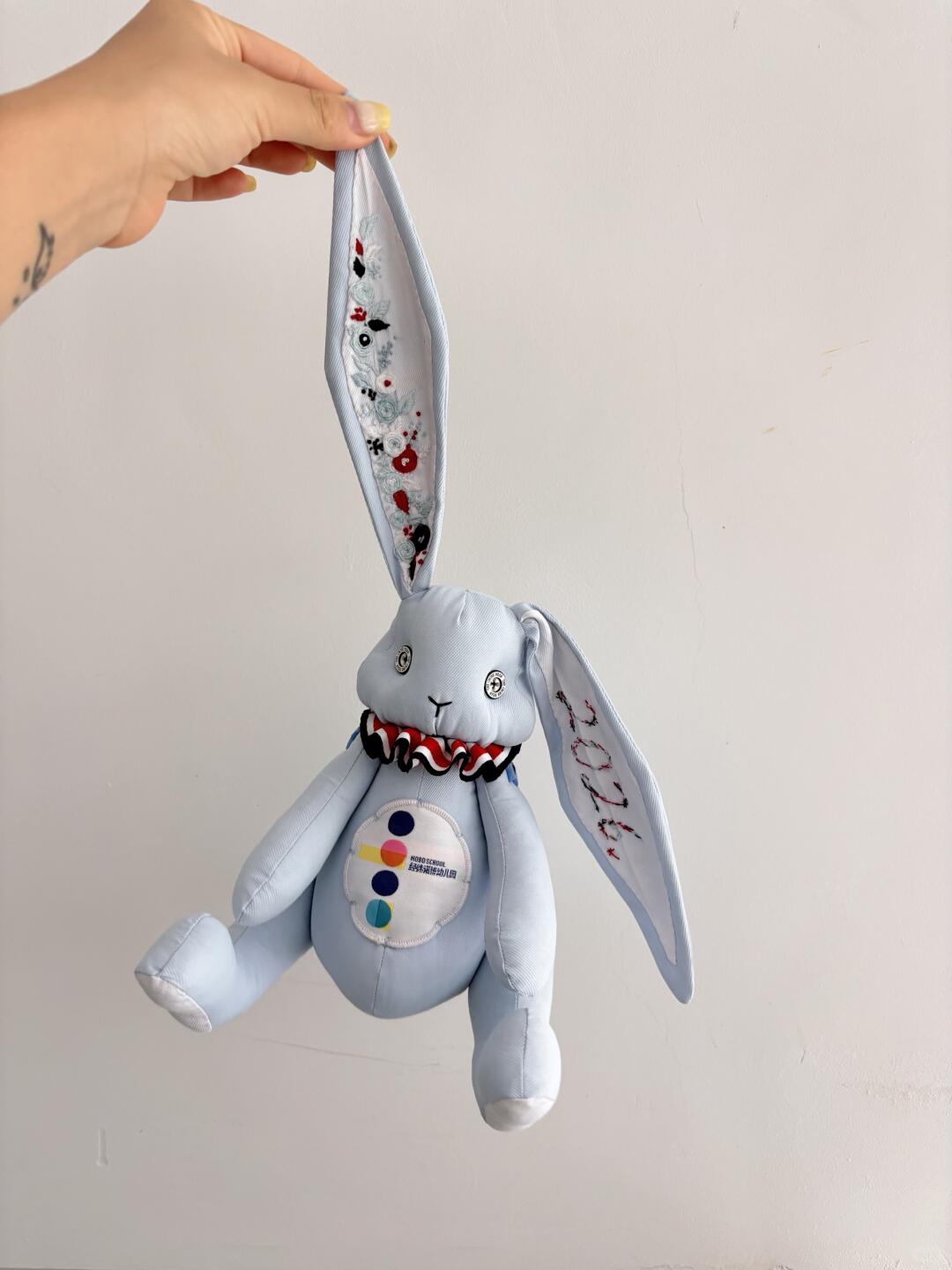 Light blue denim bunny plush hanging by one embroidered ear against a plain wall.