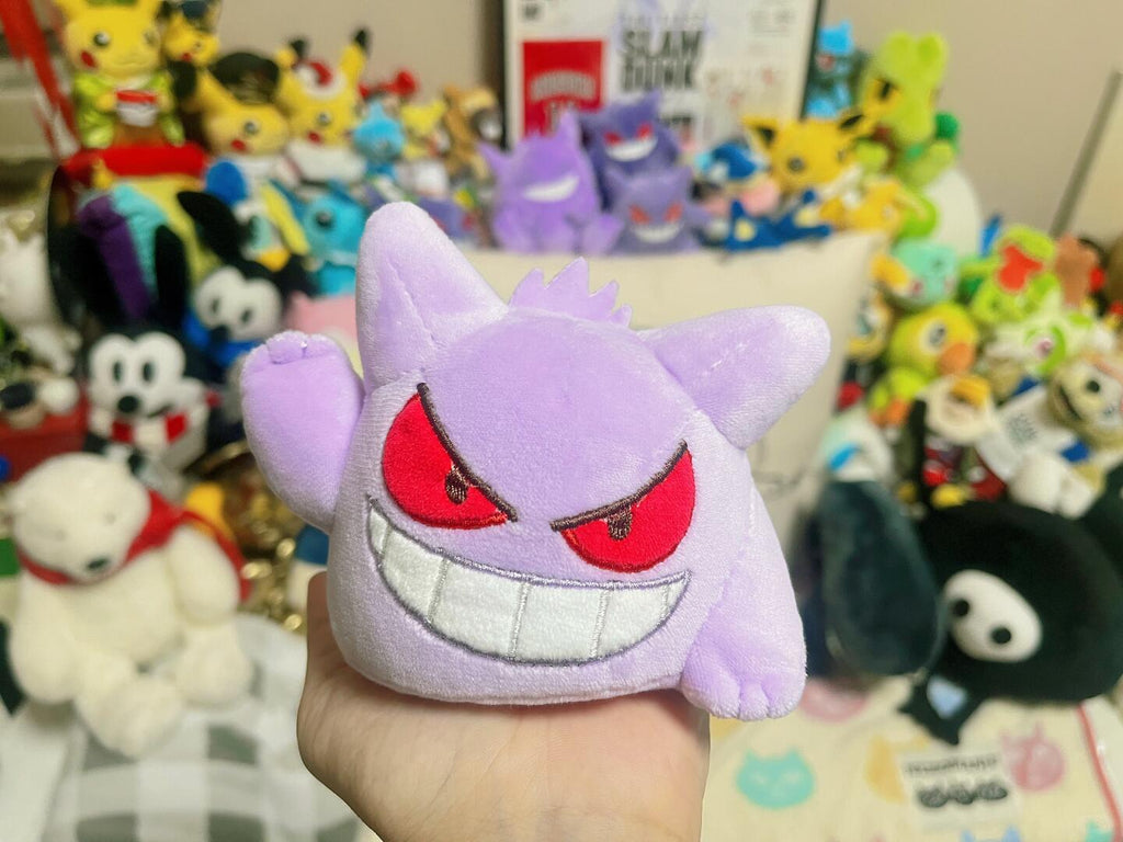 A hand holding a small Gengar purple plush toy with red embroidered eyes and a white grin, set against a busy background of various colorful plush toys and collectibles.