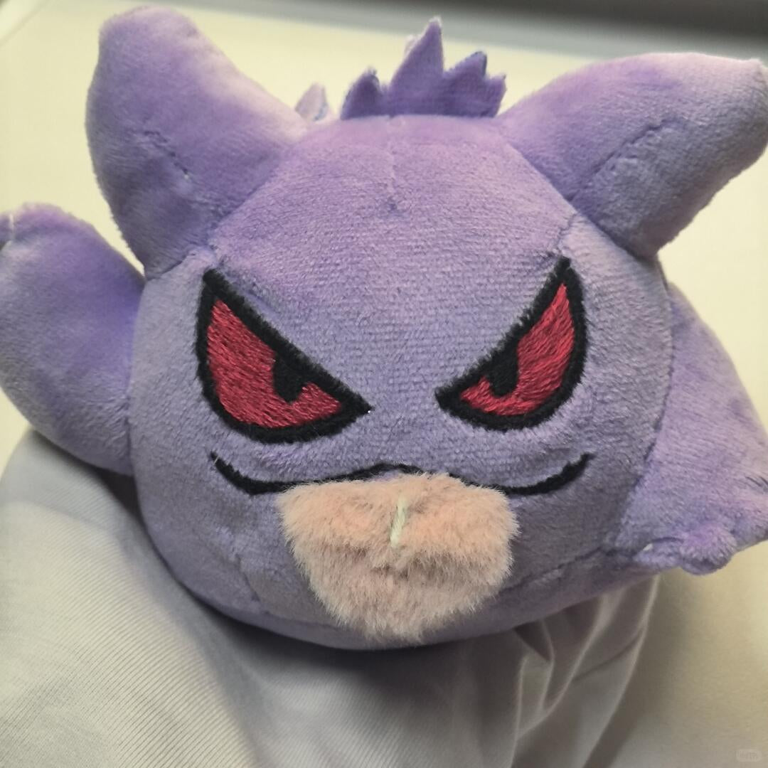A purple Gengar plush toy with red embroidered eyes and a beige muzzle, photographed close up against a light neutral background.