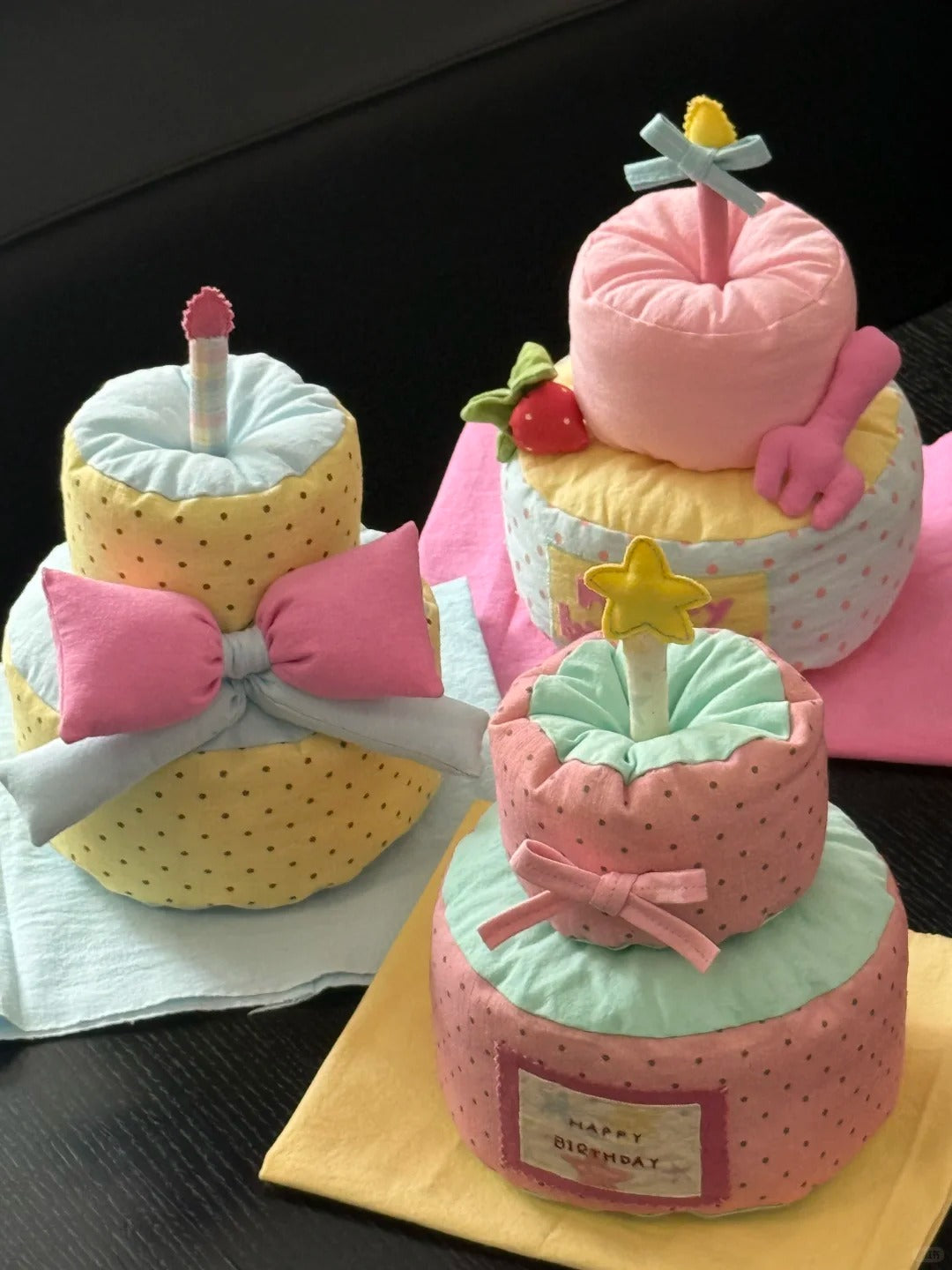 Three plush birthday cake-shaped pillows arranged on a dark tabletop, decorated with pink bows, pastel yellow and mint green fabric, star candles, and a small 'Happy Birthday' patch on the front.