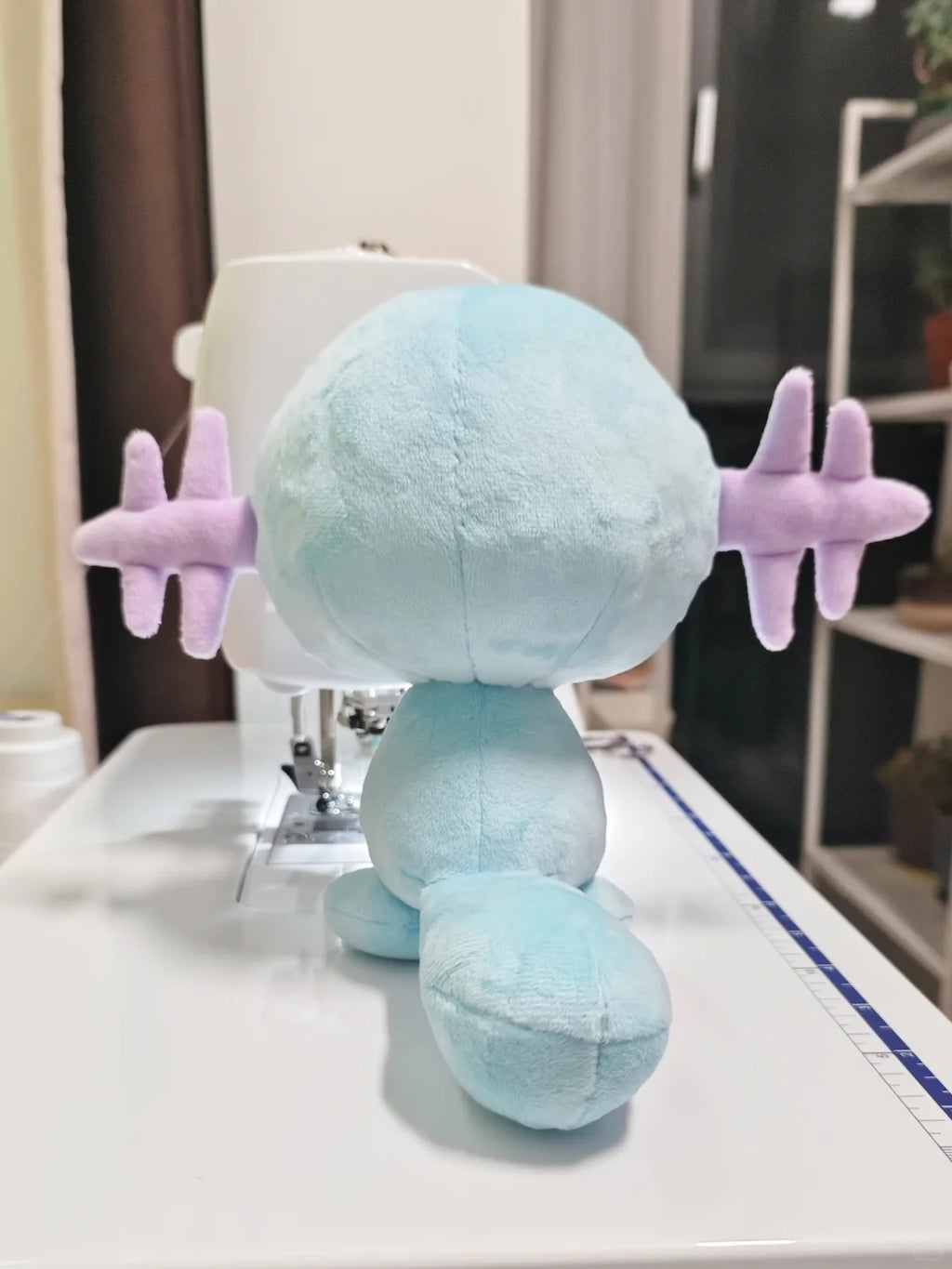 A light blue Wooper plush toy with lavender wings is shown from behind on a white sewing machine table in a home sewing room.