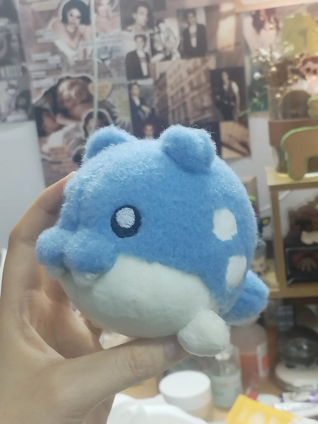 A person's hand holding a small Spheal  blue and white plush toy with a spotted pattern, set against a cluttered indoor background with posters and shelves.