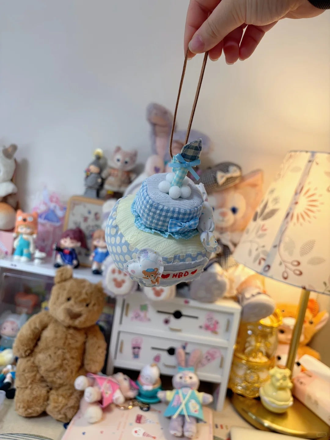 A hand holds a small blue and white diaper cake charm with a miniature diaper on top, set against a cozy, cluttered room background filled with stuffed toys, a teddy bear, and a patterned lamp.