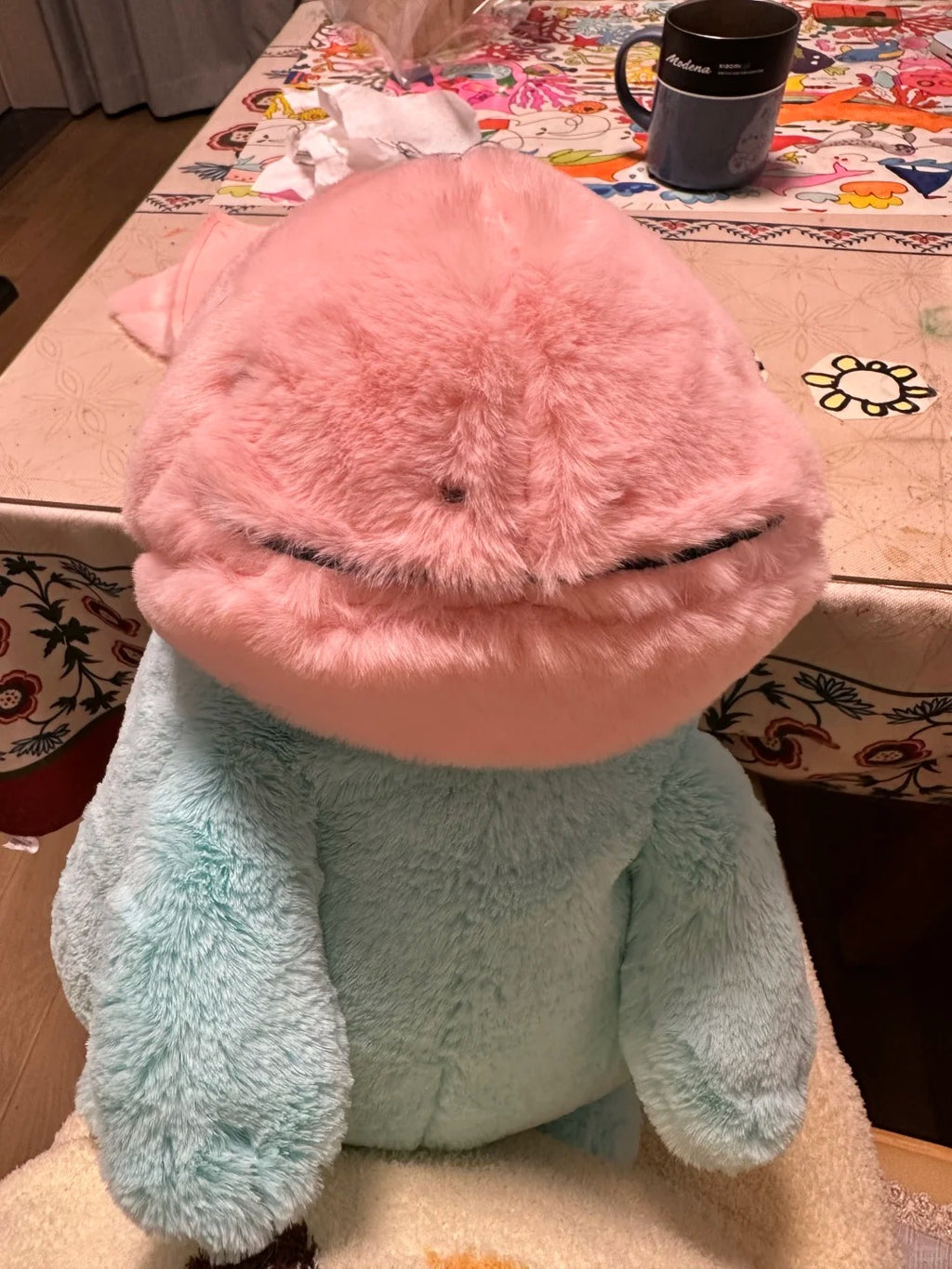 A  Quagsire plush toy with a large pink head and a light blue body, photographed indoors on a chair in front of a patterned table with a mug.