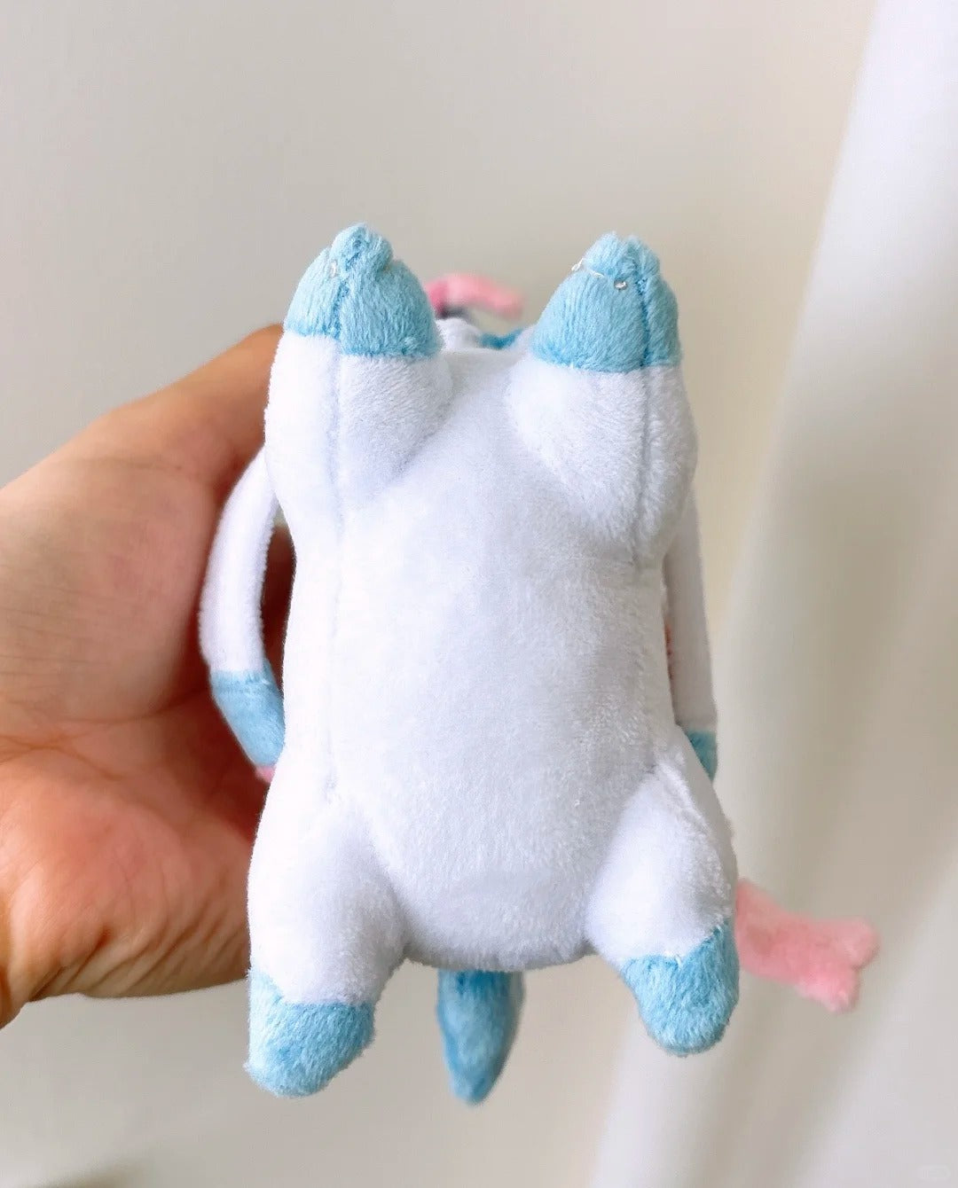 A plush toy shaped like a small white Sylveoncreature with light blue accents on its ears, paws, and tail, being held in a person's hand against a soft off-white background.