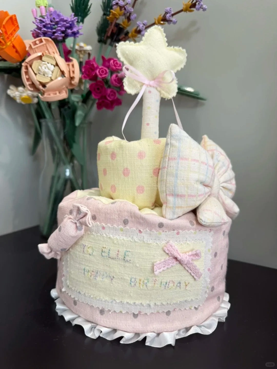 A handmade baby shower cake made from baby clothes and fabric items, decorated with a star topper, a pink bow, and a personalized name patch reading 'TO ELIE HAPPY BIRTHDAY'.