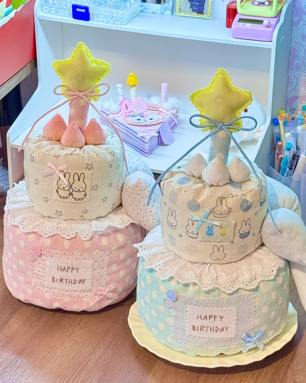 Two handmade fabric baby shower cake toppers displayed on a wooden table in front of a white toy organizer shelf. The left cake topper is pink with white polka dots, featuring a bunny patch, lace trim, and a yellow star topper.