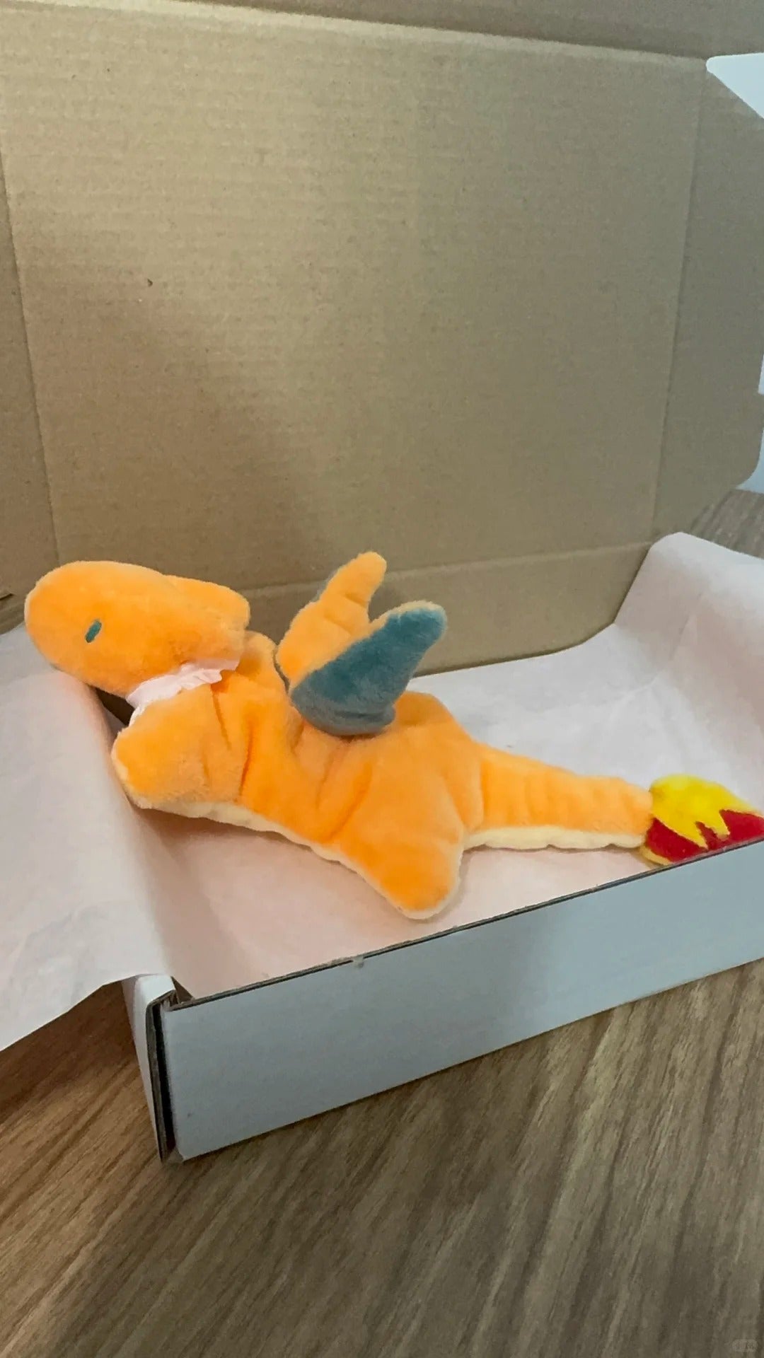 An orange dragon plush toy with blue wings and a red-yellow tail lies on white tissue paper inside an open cardboard box on a wood floor.