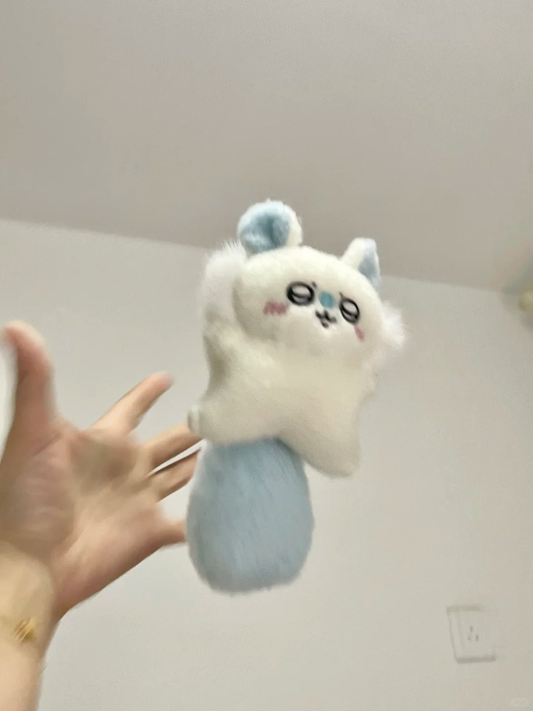 A small plush momonga accessory being held up by a person's hand against an indoor wall background. The item appears fluffy and pastel, with a white upper body, light blue ears and tail, and simple black facial details.
