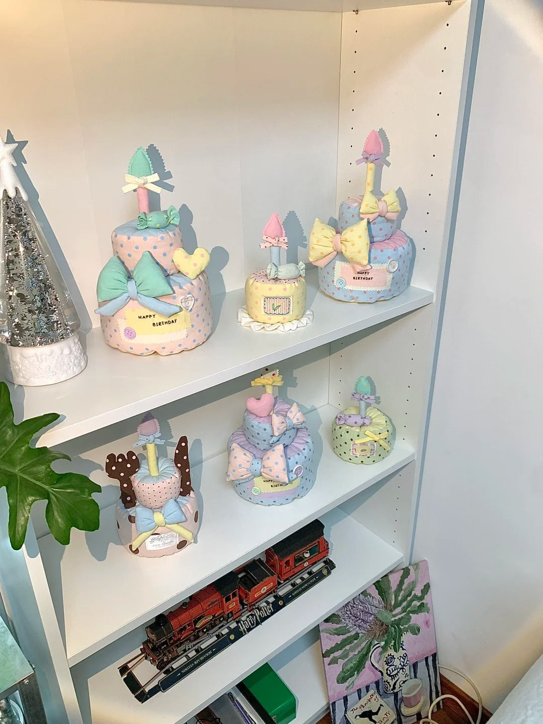 A white shelving unit displaying several decorative diaper cake toppers in pastel colors, including pink, yellow, blue, and green, alongside other household items like a toy train and a plant.