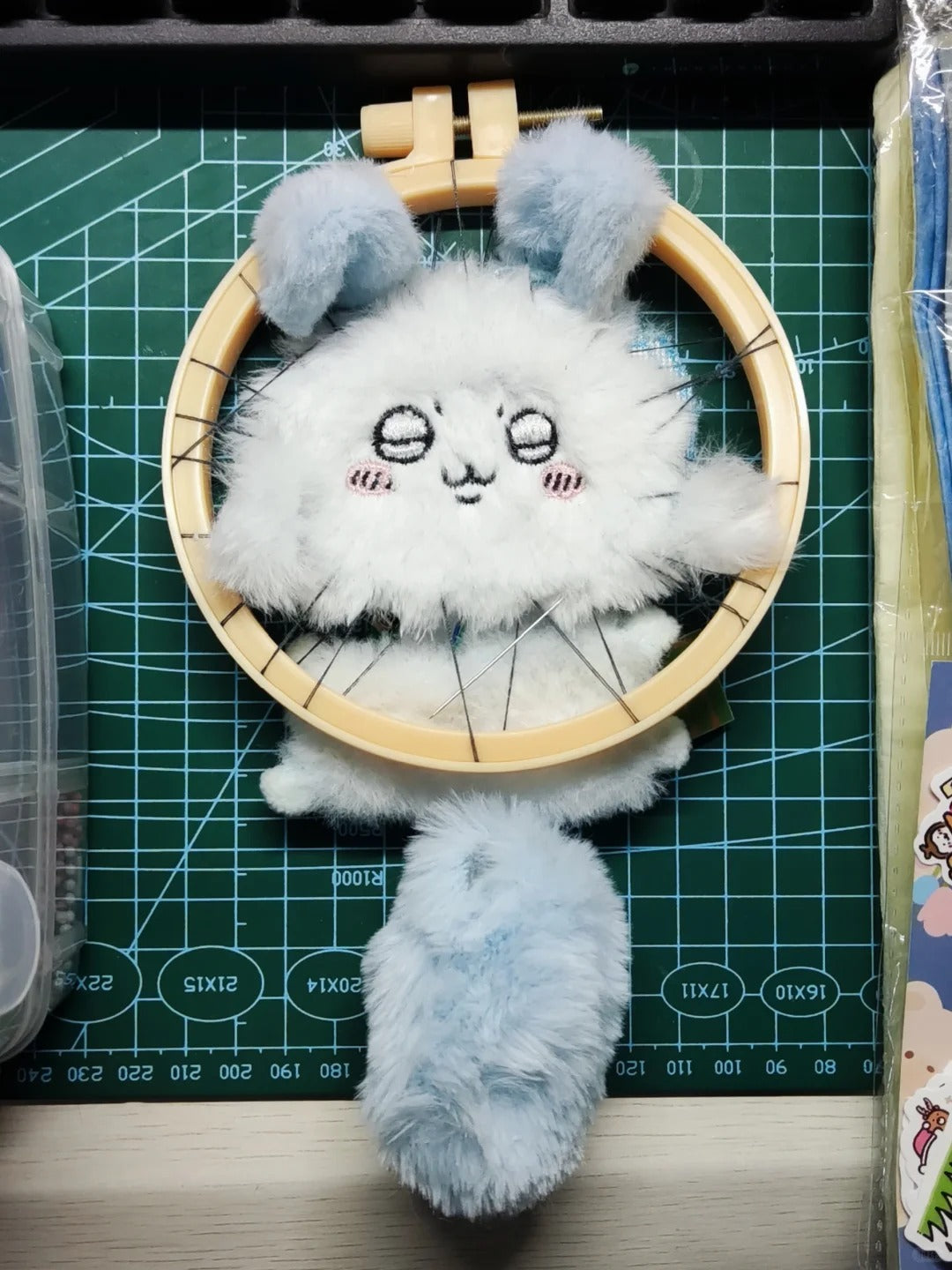 A fluffy white and light blue momonga plush character with embroidered eyes and cheeks, displayed inside a wooden embroidery hoop on a green cutting mat.