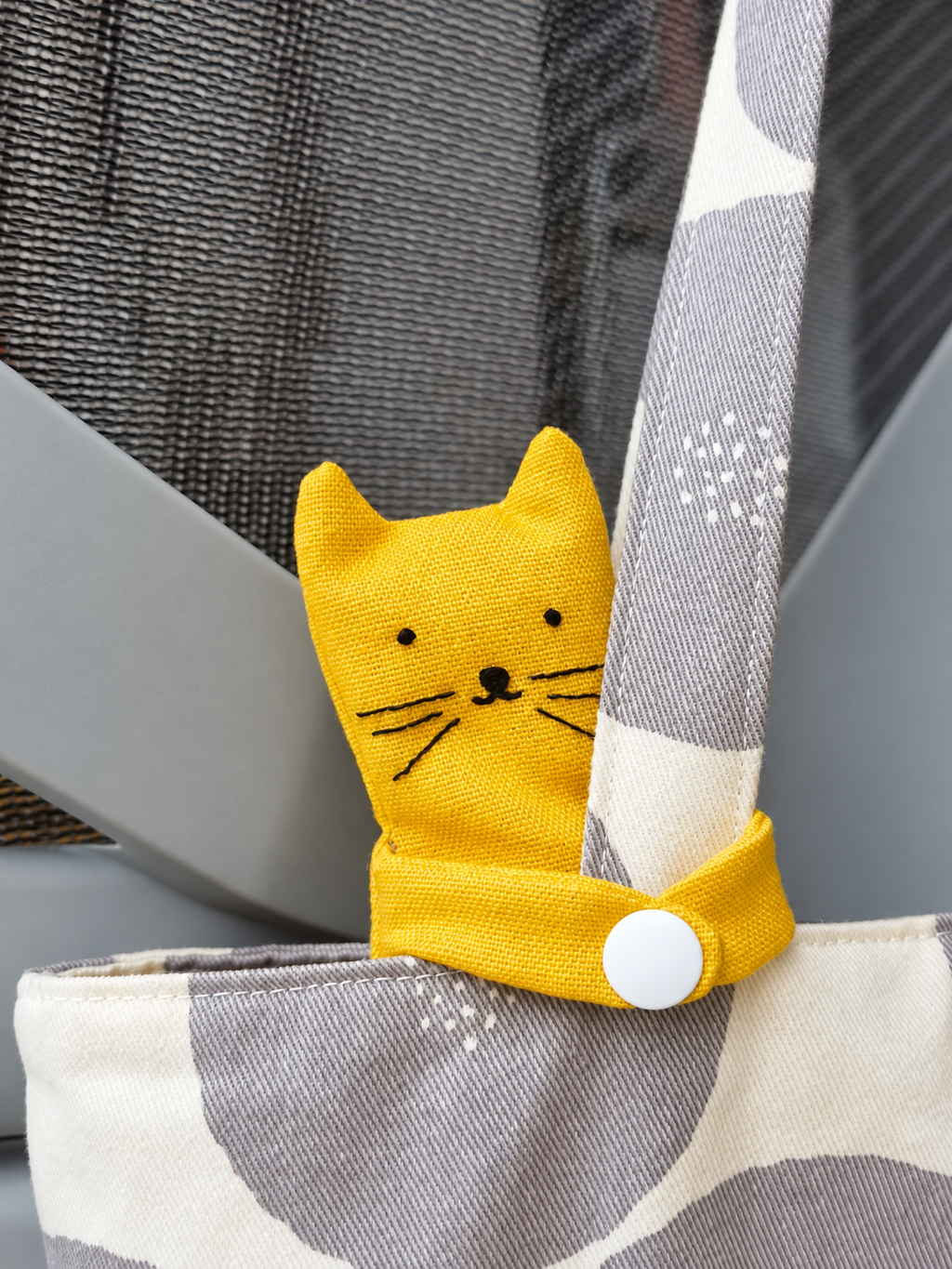 A bright yellow fabric cat-shaped bag clip with embroidered facial features and a white snap button, attached to a gray and white patterned tote bag.