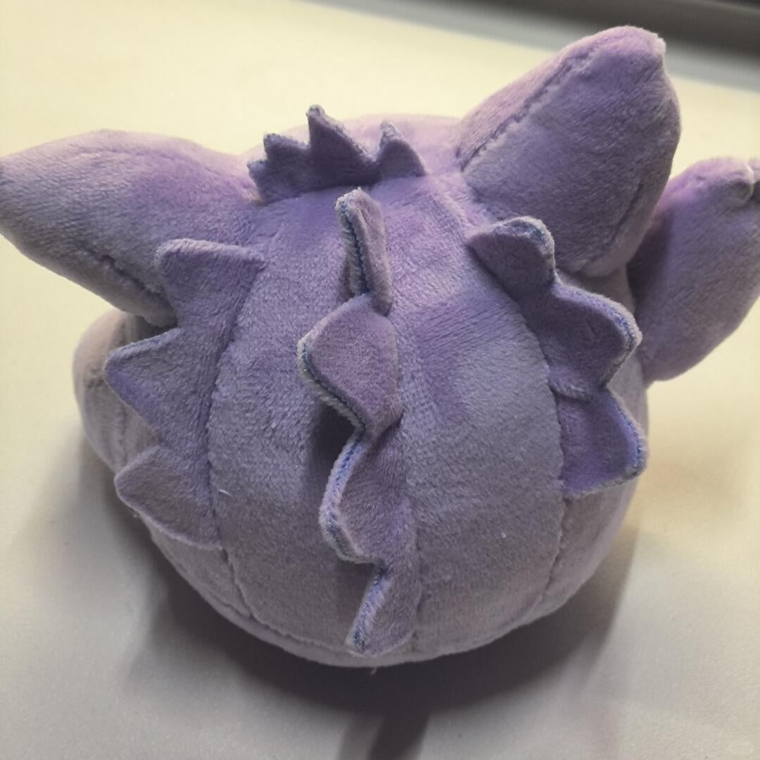 A Gengar purple plush toy with a rounded body, spiky back, and pointed ears, photographed on a plain light surface.