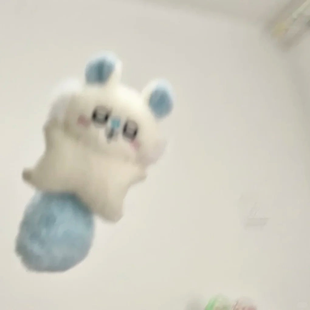 A small momonga plush toy with a white body, light blue ears, and a light blue tail, shown against a plain off-white background. The image is noticeably blurry and out of focus.