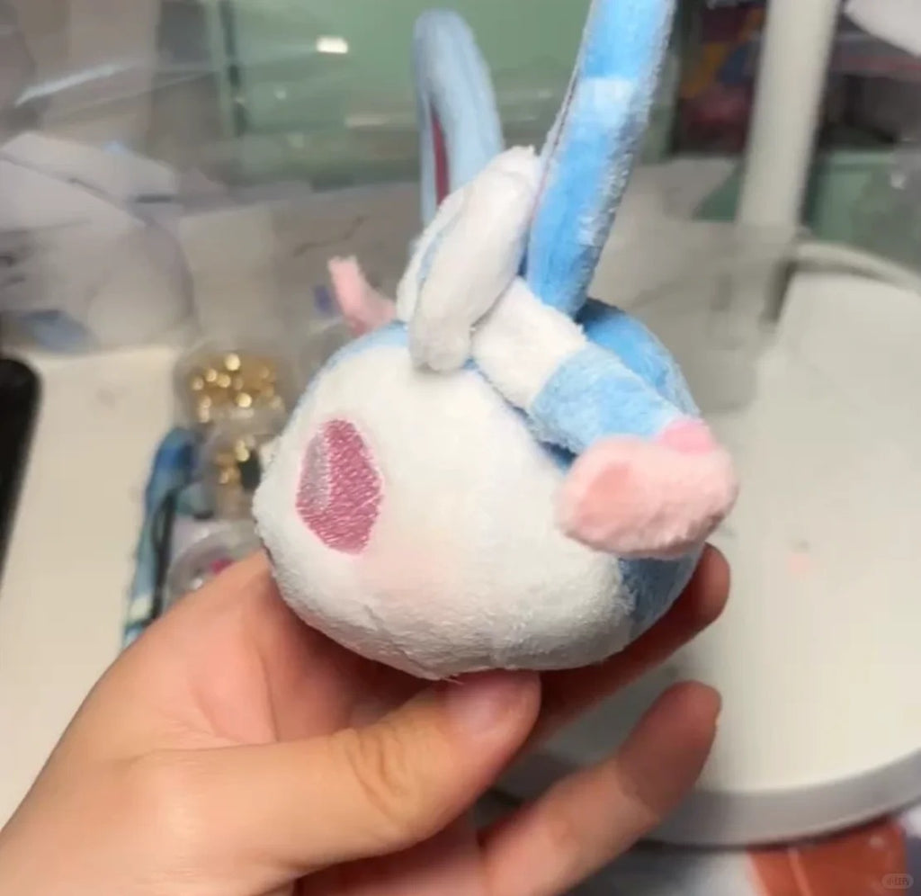 A hand holding a small  Sylveonplush toy with a blue and white striped body, pink heart-shaped face, and long blue ears, photographed in a casual indoor setting.