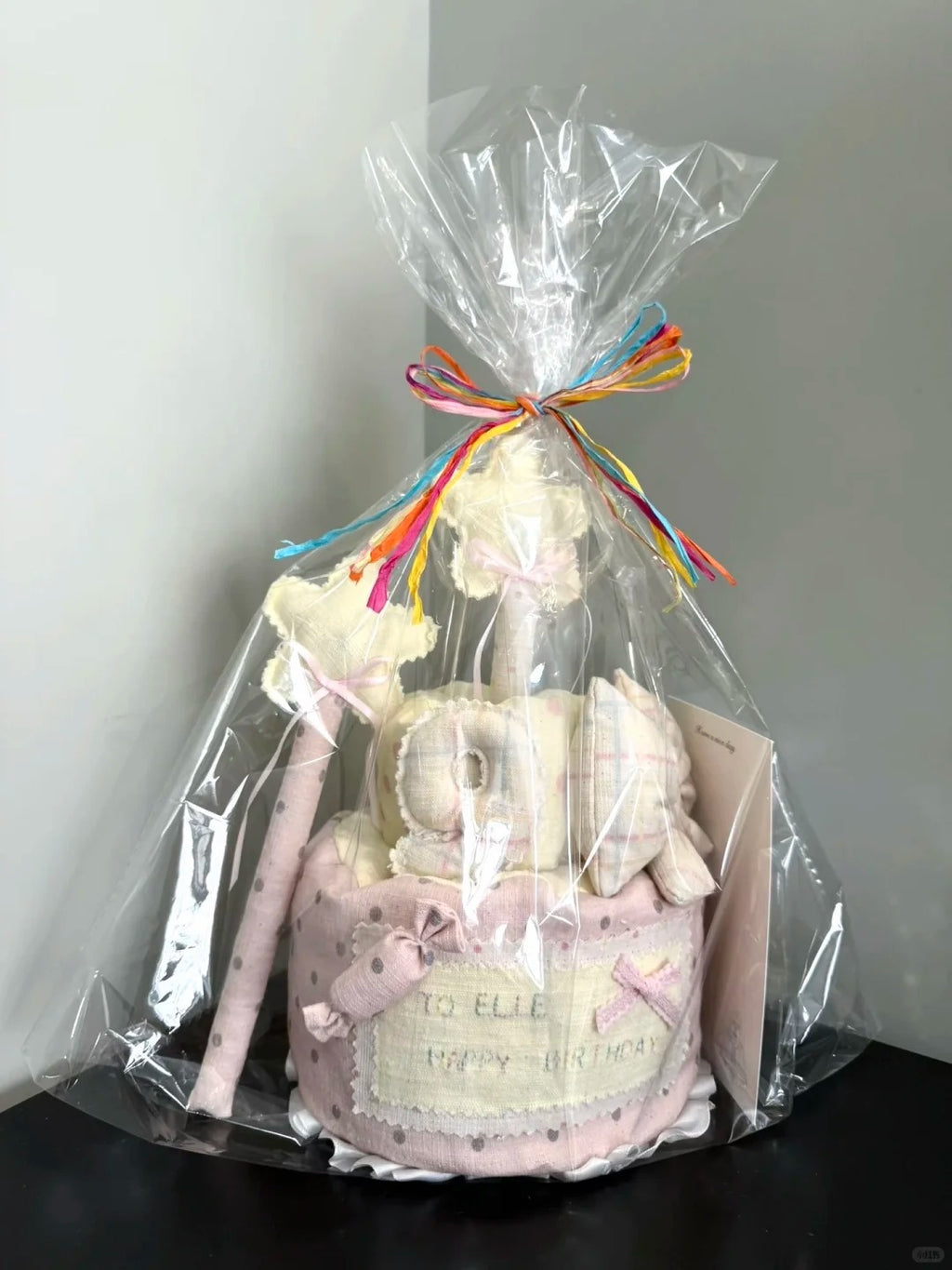 A baby shower diaper cake gift set wrapped in clear cellophane and tied with a rainbow-colored bow, sitting on a dark surface against a light gray wall.