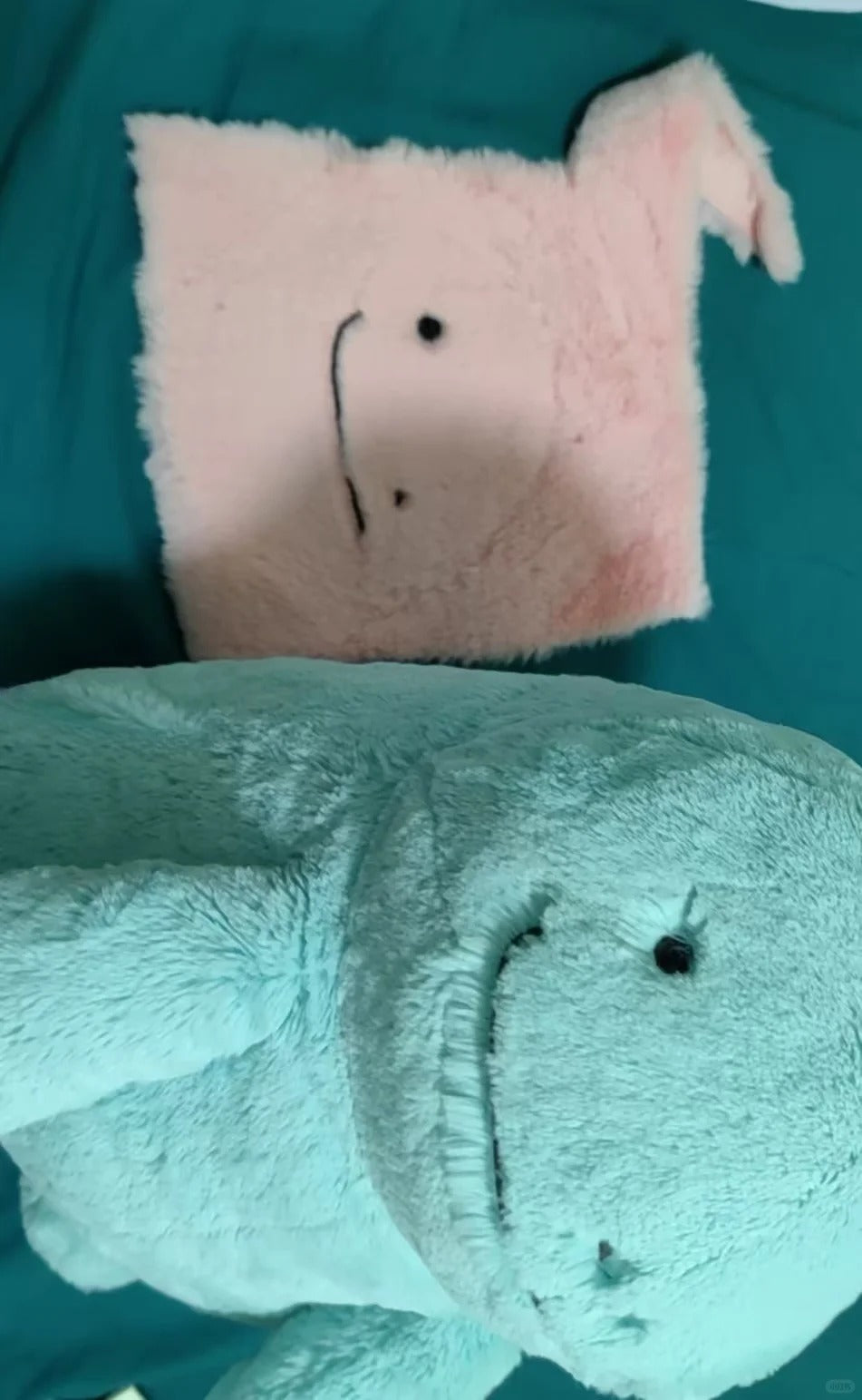 A light blue plush Quagsire toy in the foreground and a small pink plush pillow with a simple stitched face in the background, both placed on a teal fabric surface.