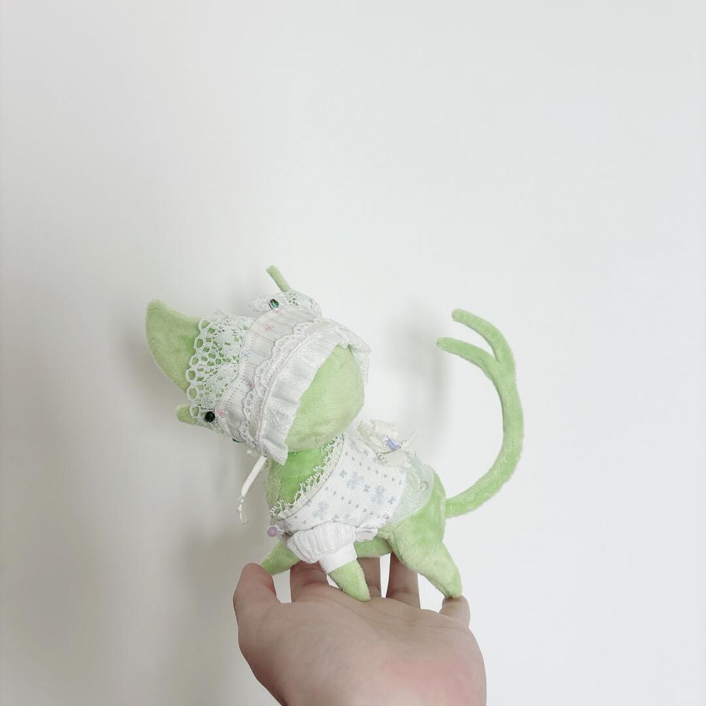 A small light green plush cat wearing a white lace-trim pet dress, held in a person's hand against a plain white background.