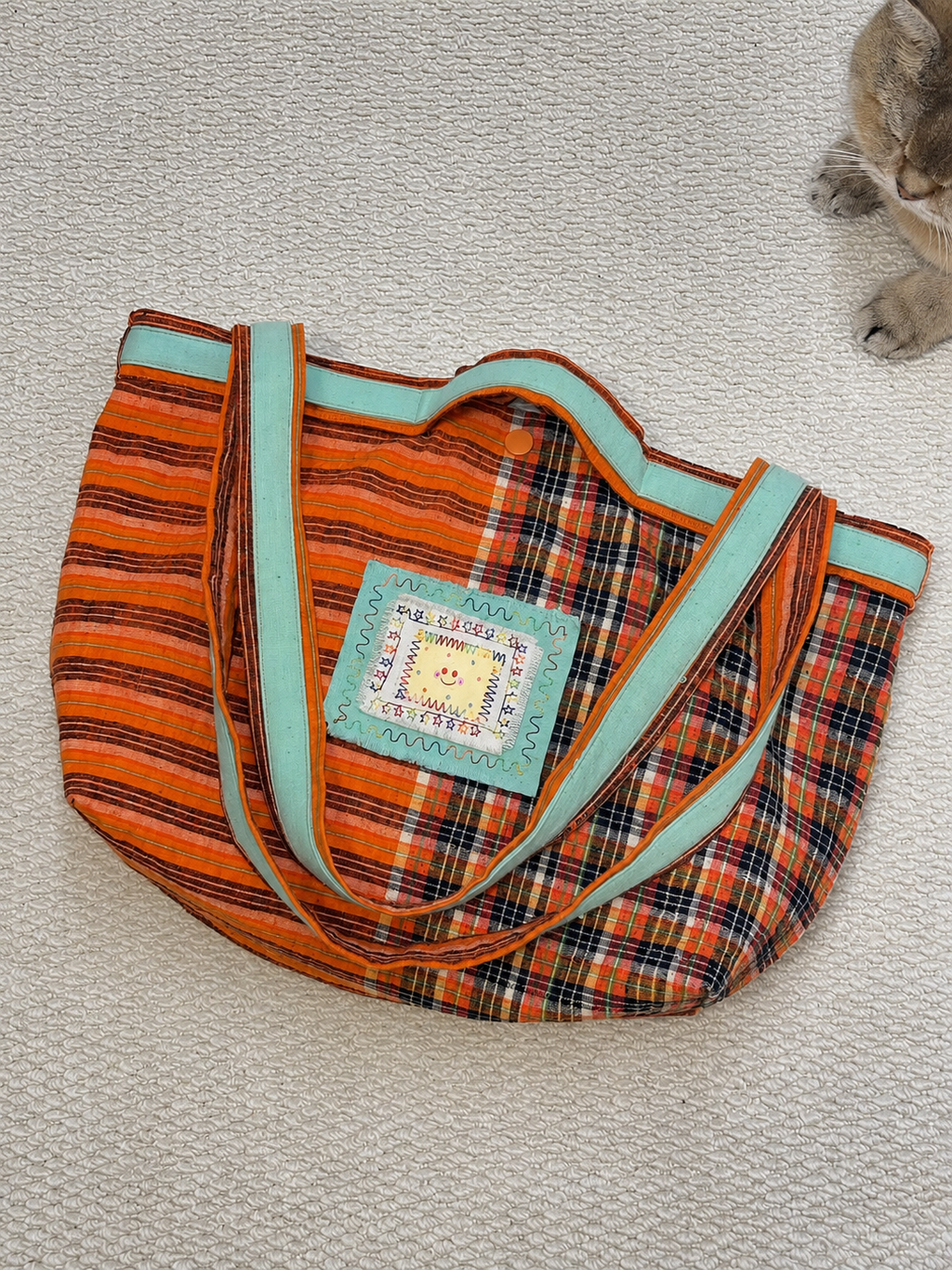 A handmade fabric tote bag with orange, black, and teal stripes, featuring a central decorative patch with a smiling sun and the text 'Happy Birthday!'. A cat's paws are visible in the top right corner on a textured beige carpet.