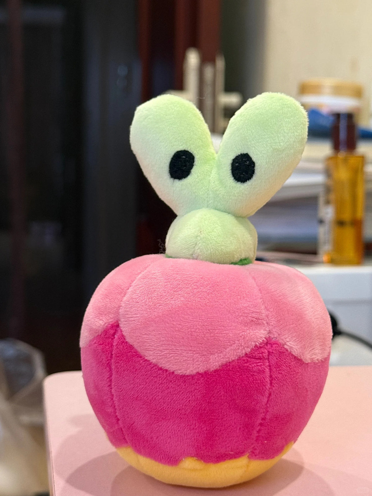 A Applin plush toy shaped like a pink apple with a light green cartoon face and black eyes emerging from the top, set on a pink surface with a blurred indoor background.