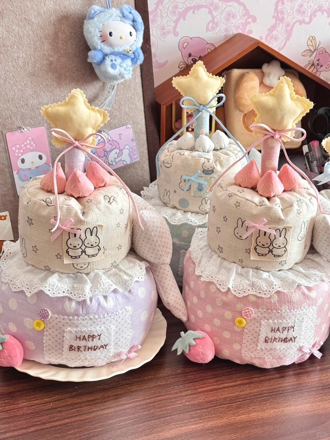 Three decorative fabric cake-shaped baby shower favors or gifts arranged on a wooden table, featuring pastel pink, lavender, and cream colors with bunny prints, lace trim, star toppers, and strawberry accents. A Hello Kitty plush hangs in the background, and a small house decor piece is visible behind the items.