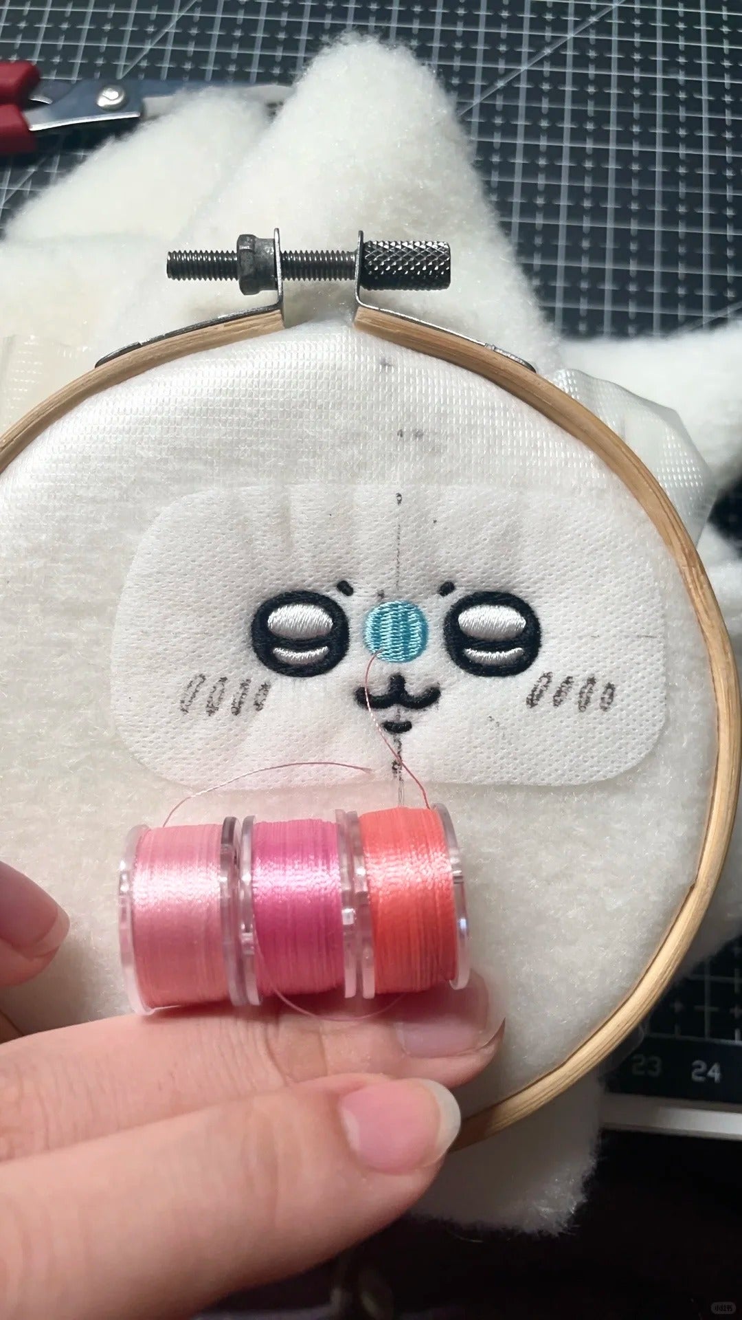 A close-up of a hand holding a wooden embroidery hoop with white fabric and a partially finished embroidered face. A hand is holding two spools of thread, one pink and one coral, in front of the hoop.