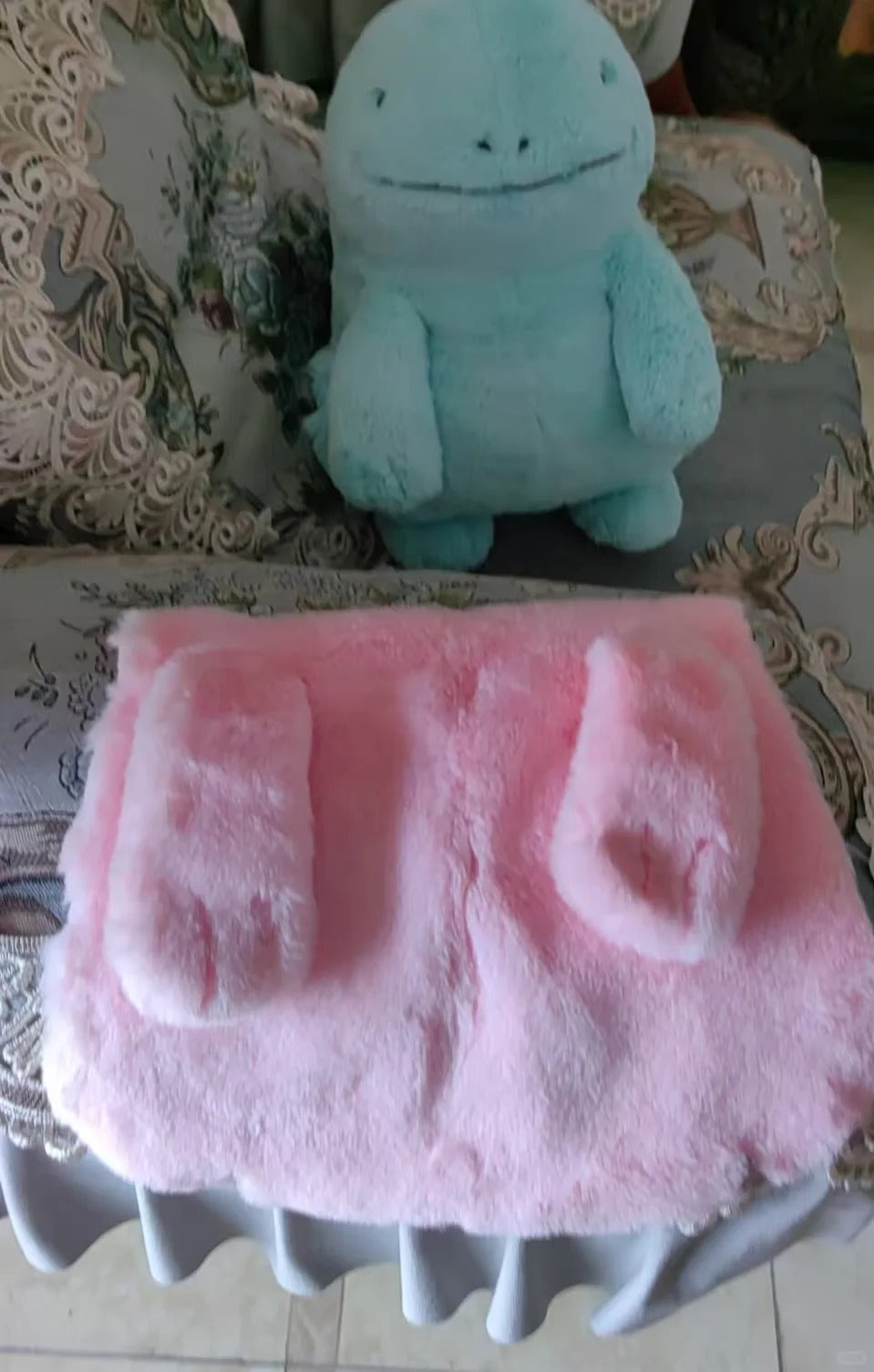 A pink plush Quagsire belly with two matching bootie-shaped pieces laid flat in front of a light blue stuffed toy on a patterned sofa.