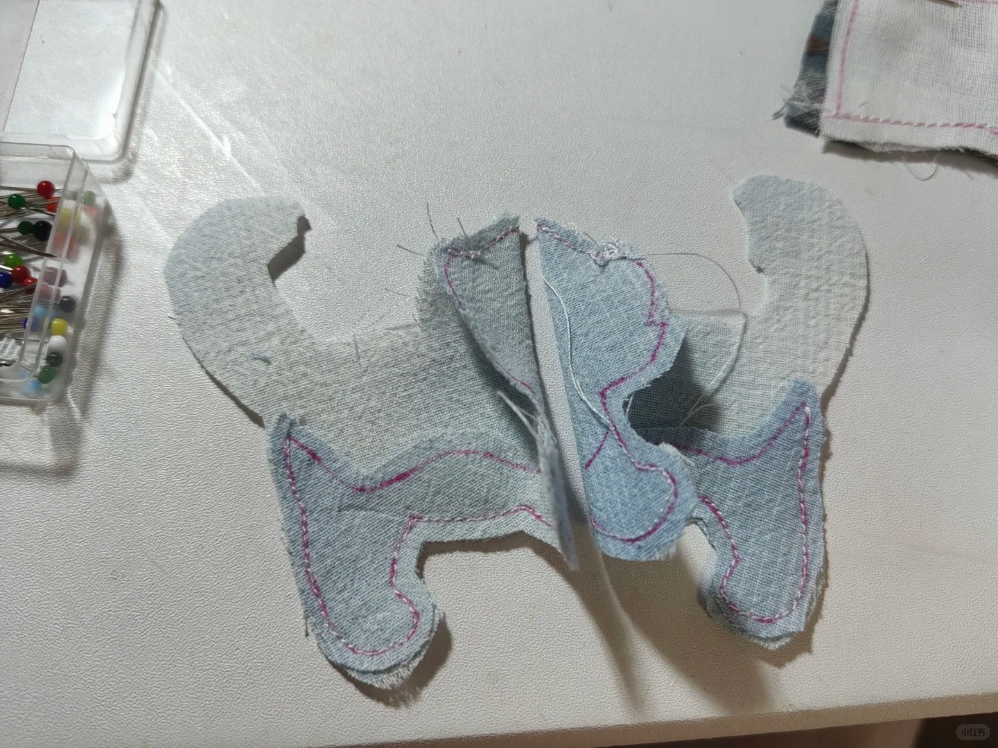 A butterfly-shaped fabric patch or applique made from light blue and white textured fabric with pink stitched outlines, placed on a white tabletop with sewing supplies nearby.
