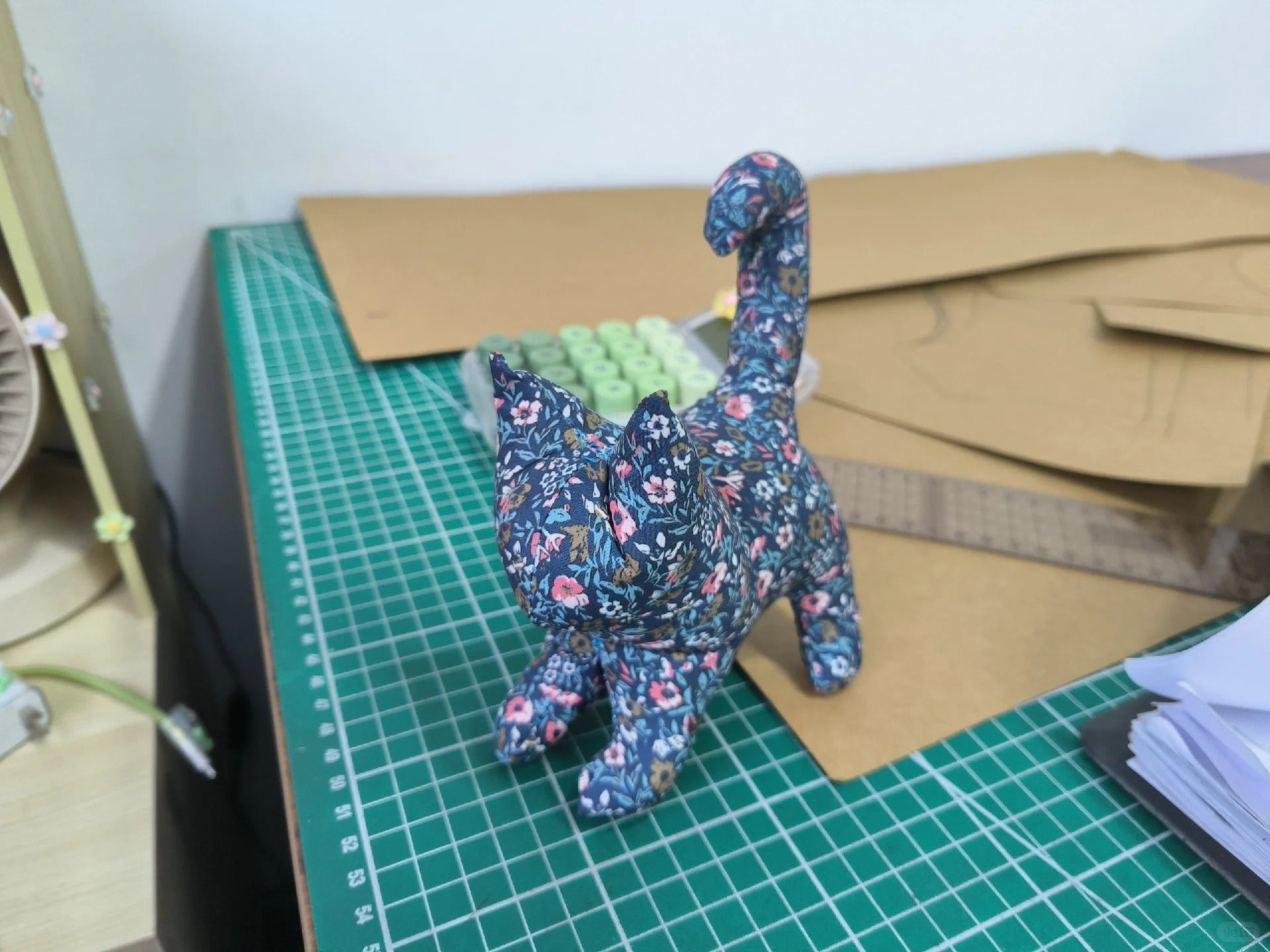 A small handmade cat-shaped fabric ornament or figurine with a blue floral print, standing on a green cutting mat on a worktable with cardboard templates and sewing supplies in the background.